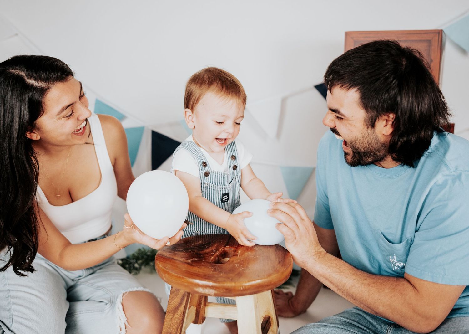 Joyful family portrait with baby boy on a wooden stool holding white balloons at birthday shoot.