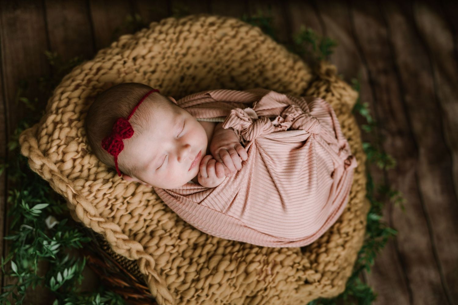 Sleeping newborn baby girl in striped pink swaddle with red bow headband in knit basket with greenery.