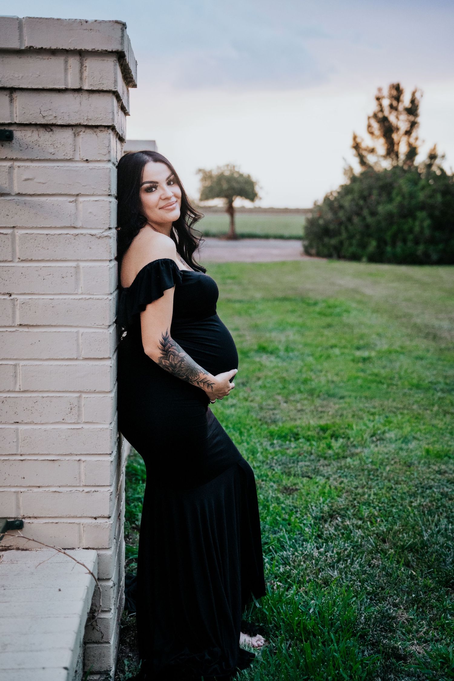 Pregnant woman in elegant black off-shoulder dress leaning against brick wall, holding baby bump with tattooed arm.