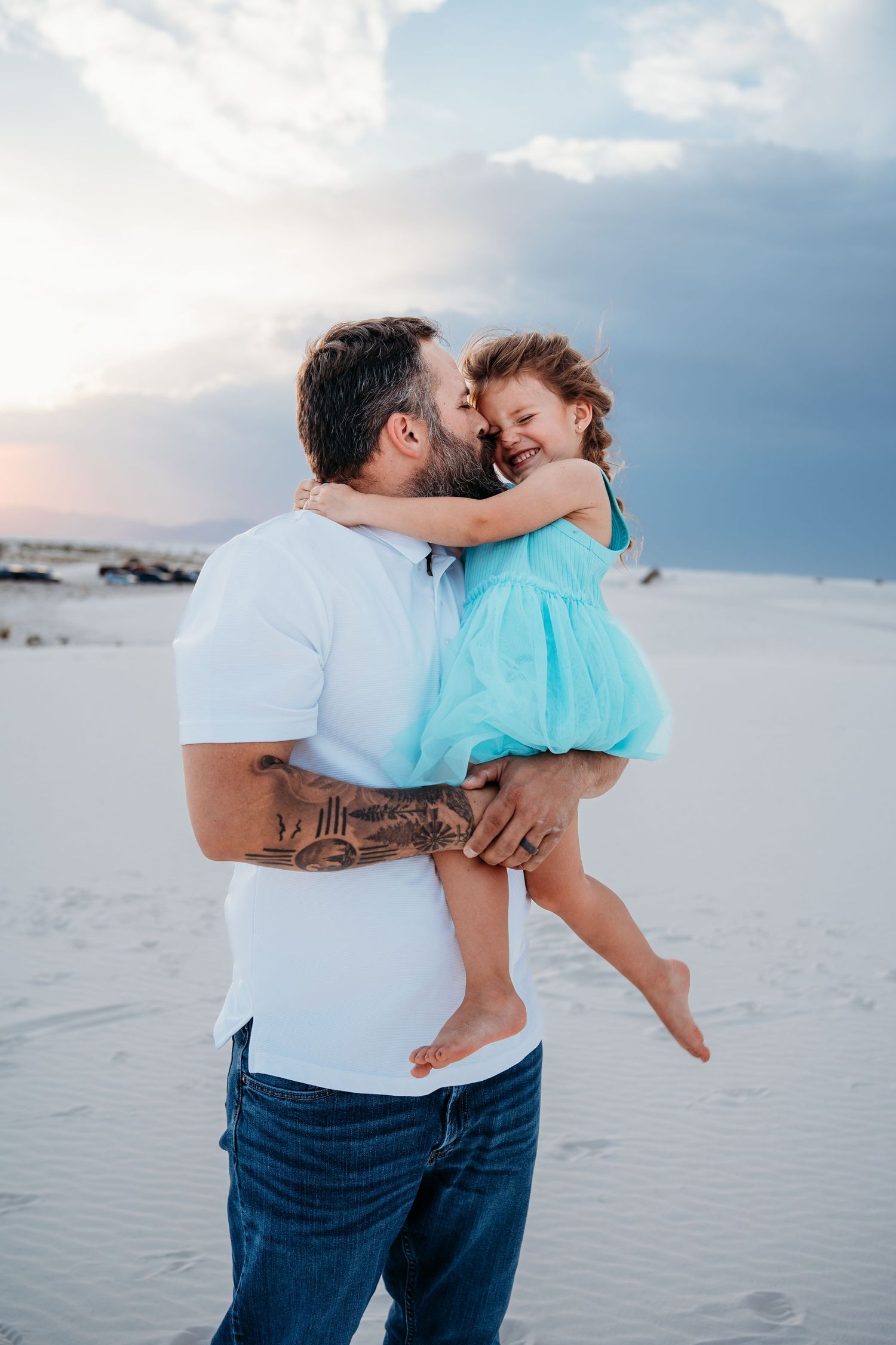 Tattooed father holding laughing daughter in turquoise dress close on white sand dunes at dusk.