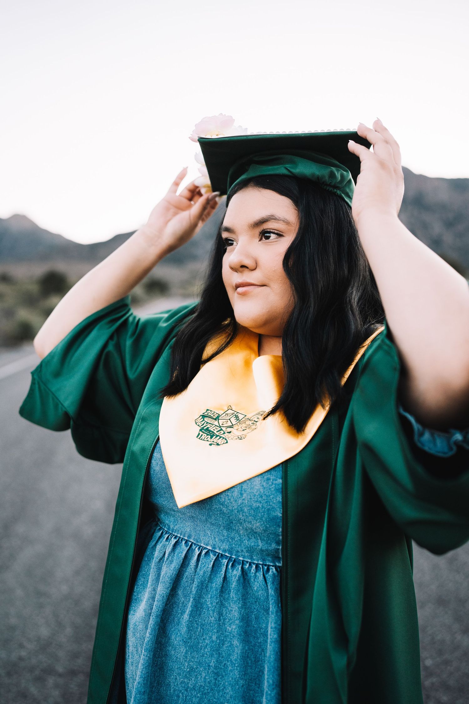 Young woman in green graduation cap and gown adjusting her cap outdoors with mountains in background.