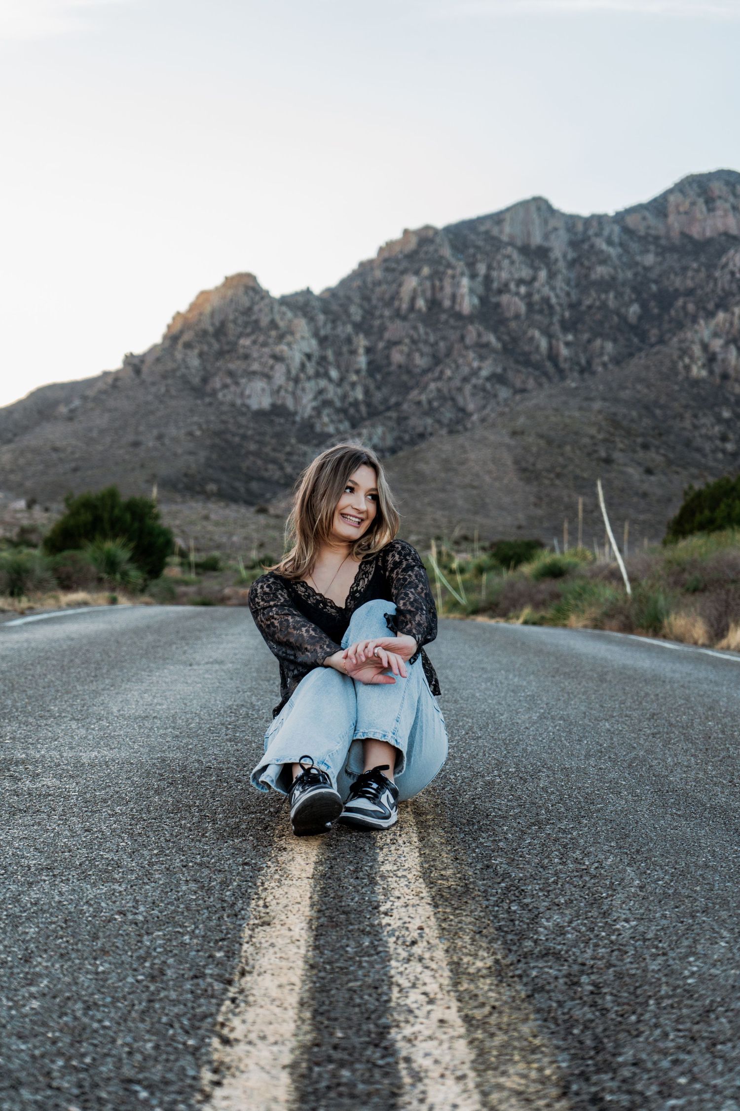 Cheerful young woman in black lace top sits cross-legged on desert highway with mountain views.