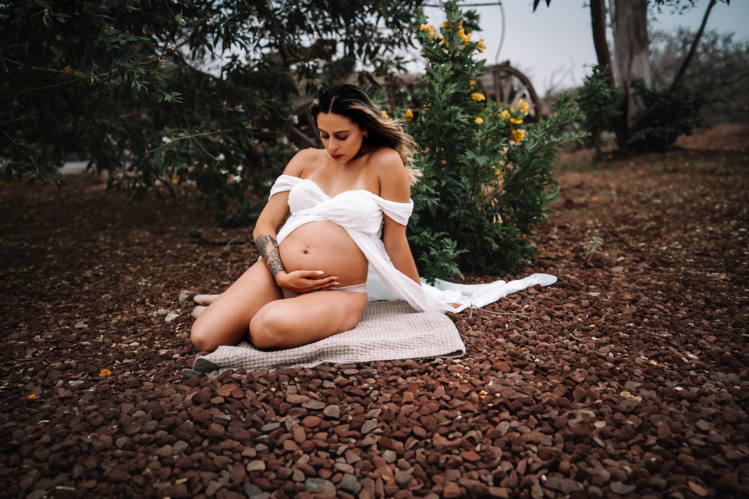 Pregnant woman in white off-shoulder outfit sits on white cloth amid brown pebbles in outdoor garden.