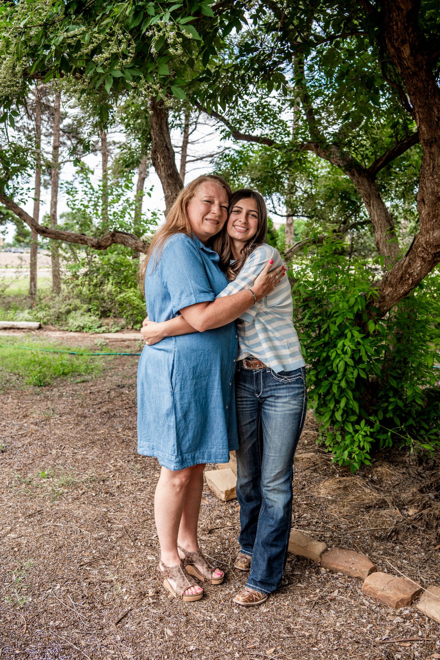 Two women share a warm embrace while smiling under a leafy tree in an outdoor setting.