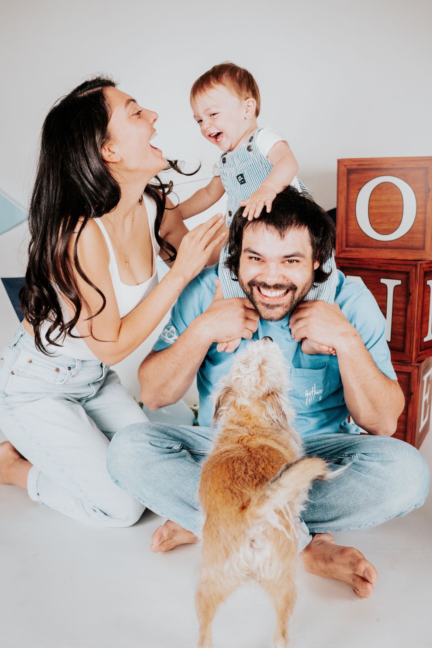 Joyful family portrait with laughing baby on dad's shoulders, smiling mom, and a golden dog in white studio.