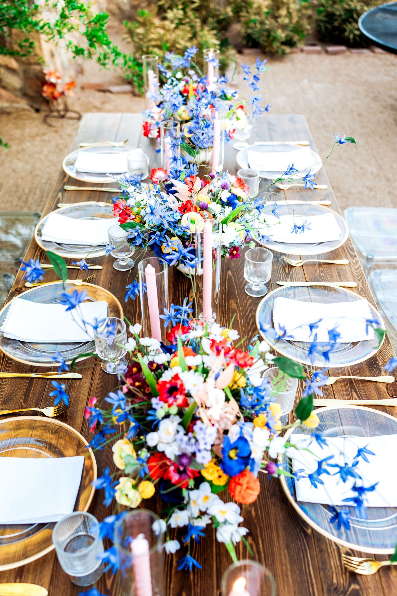 Overhead view of rustic wooden outdoor dining table with colorful wildflower centerpieces and gold cutlery.
