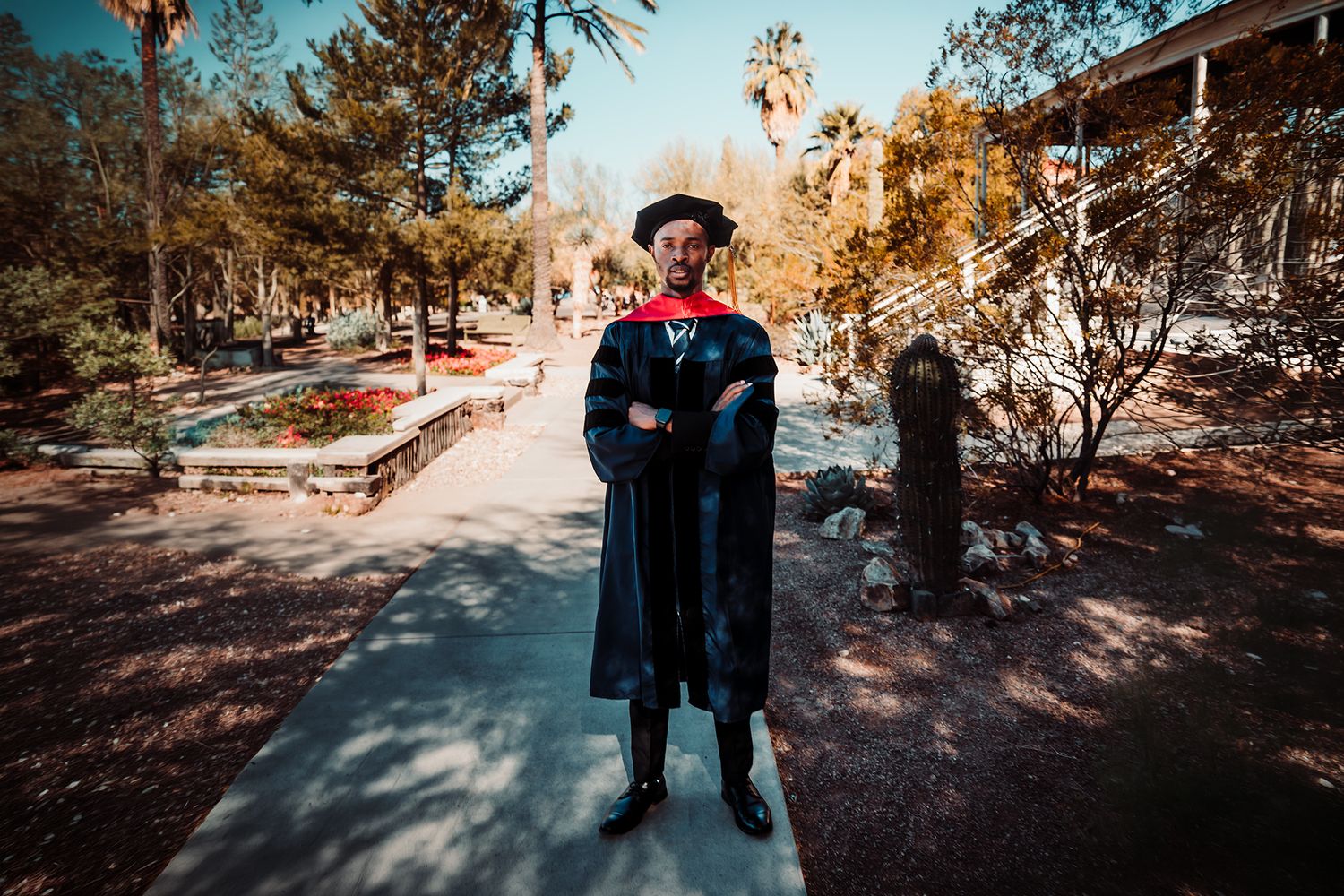 Graduate in black cap and gown stands confidently on a tree-lined campus pathway during graduation day.