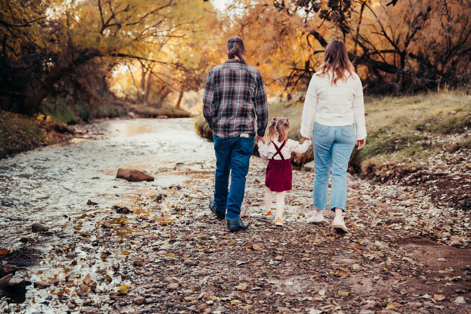 Family of three walking hand-in-hand along a rocky creek path surrounded by fall foliage.