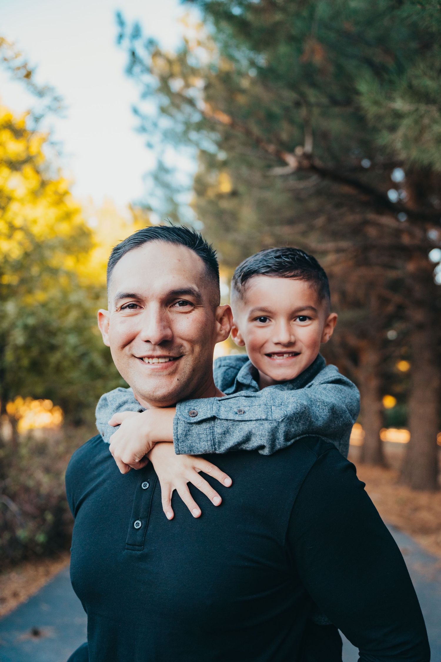 A smiling father giving his young son a piggyback ride along a tree-lined park pathway at golden hour.