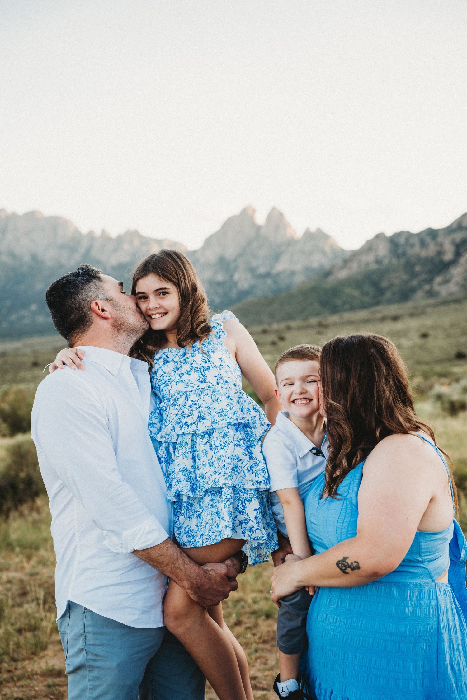 Father kissing daughters cheek while family laughs together in front of scenic mountain landscape.