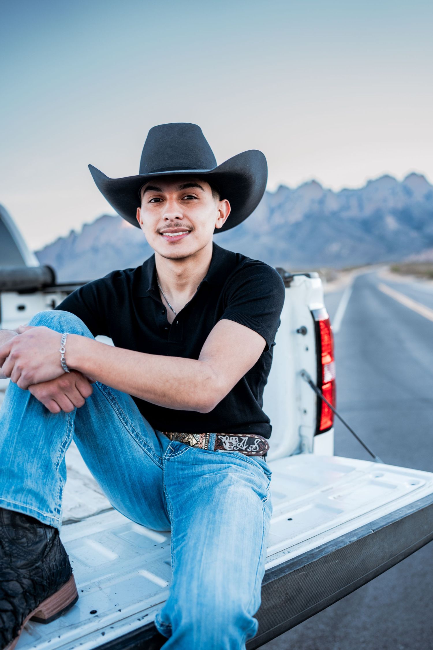 Smiling young man in black cowboy hat sits on white truck tailgate on open desert highway.