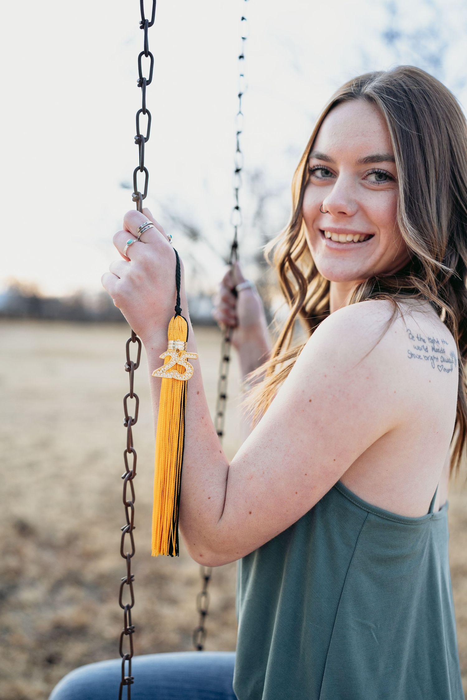 Smiling senior girl sits on chain swing displaying class of 25 graduation tassel and shoulder tattoo.