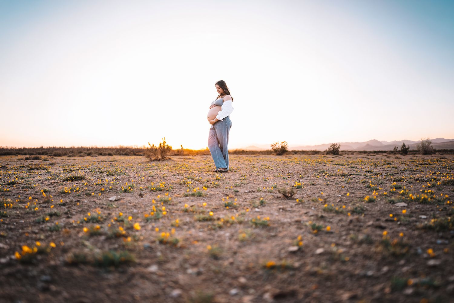 Pregnant woman standing in desert field holding baby bump during stunning golden sunset.