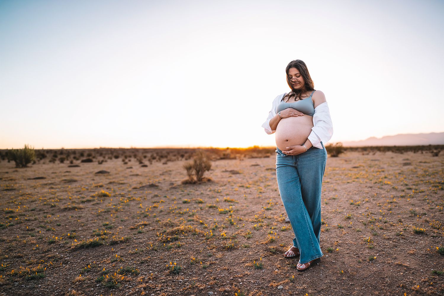 Pregnant woman in jeans and open white shirt holding bare belly in desert sunset.