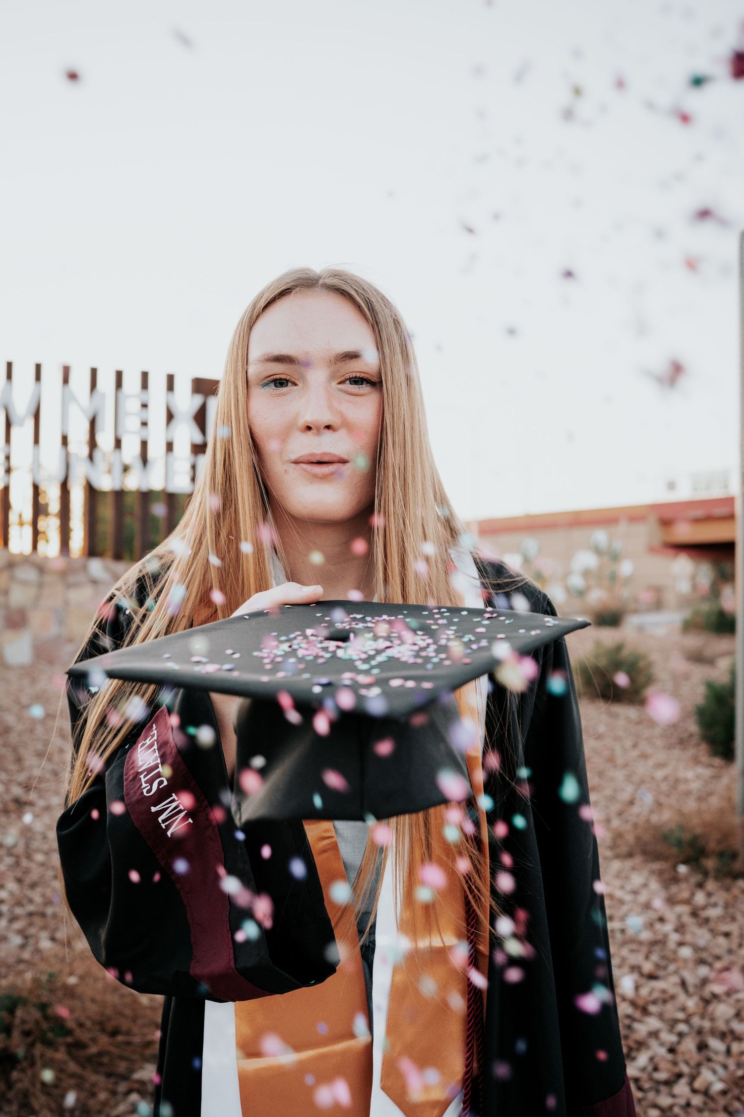 Blonde graduate holds decorated cap while confetti falls during outdoor graduation celebration.