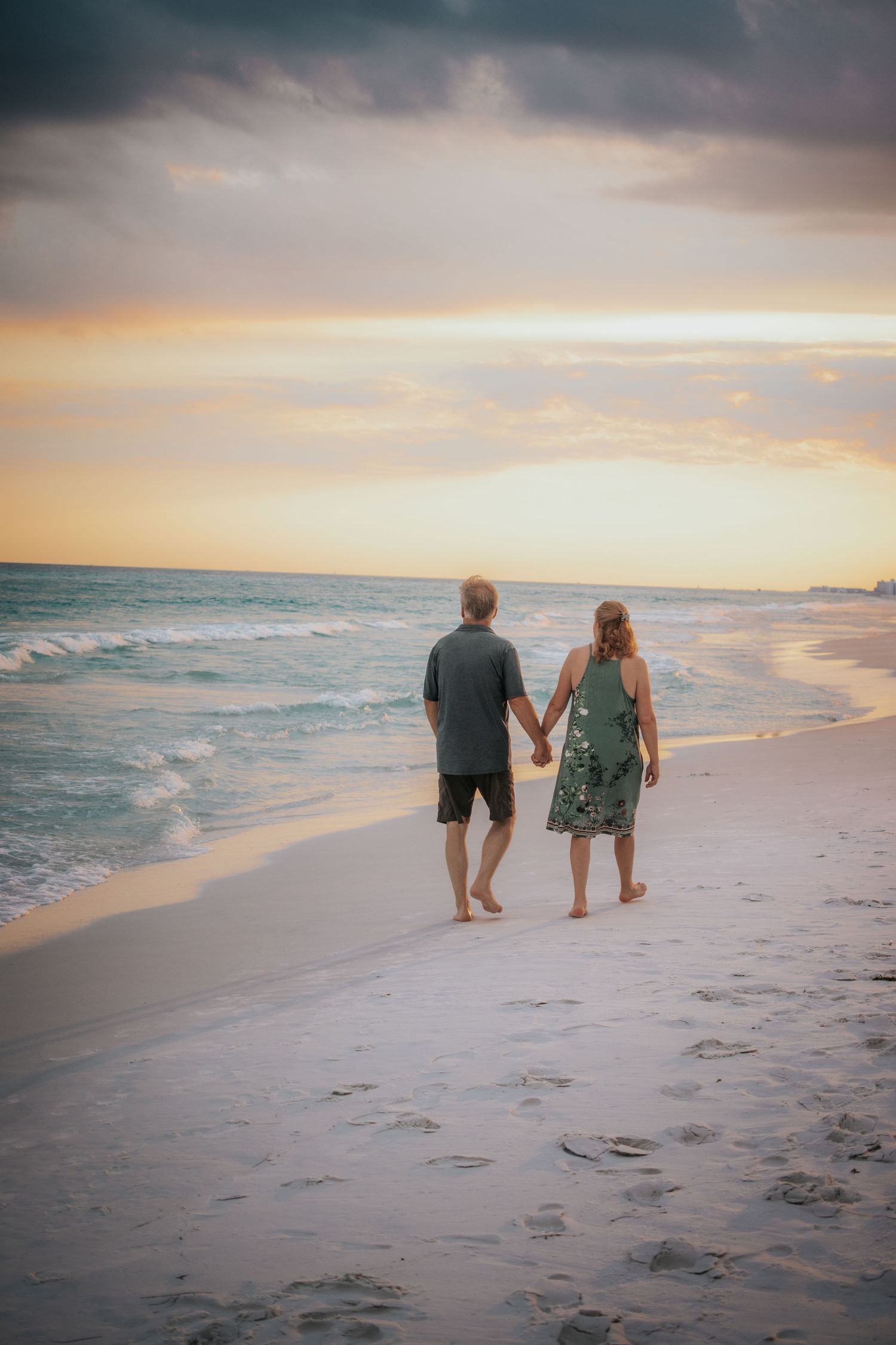 Couple walking hand in hand along a sandy beach at a colorful sunset by the ocean.