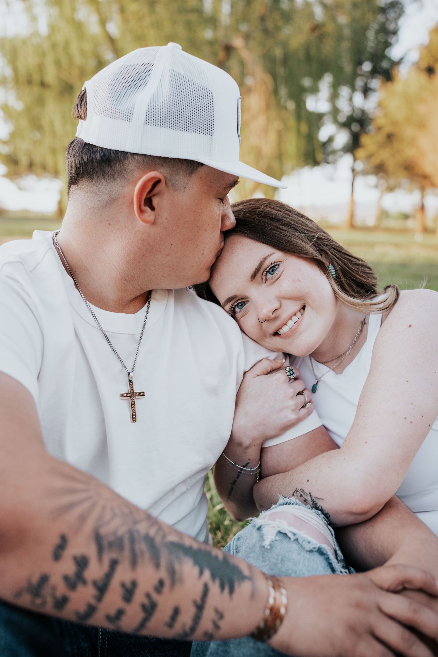 Man in white trucker cap kissing smiling blue-eyed woman on cheek during golden hour outdoor photo session.