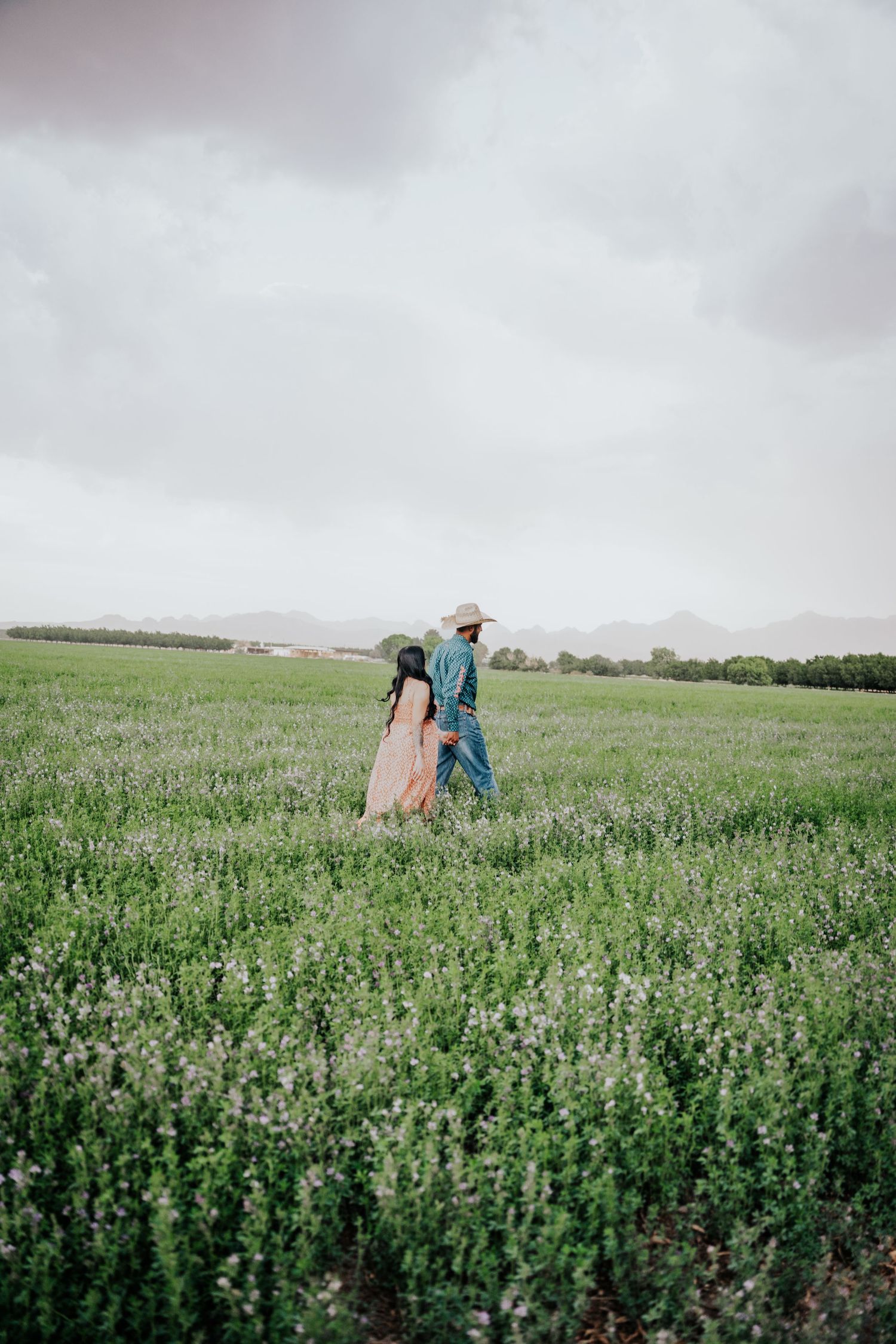 Couple walking hand in hand through a vast green field with mountains visible in the cloudy background.