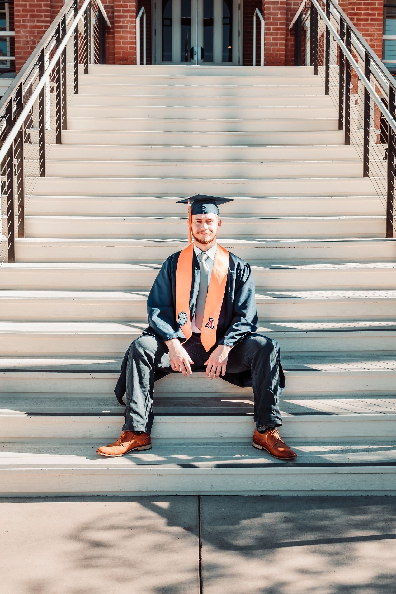 Young man in graduation cap and gown with orange stole sits proudly on university steps for graduation photo.