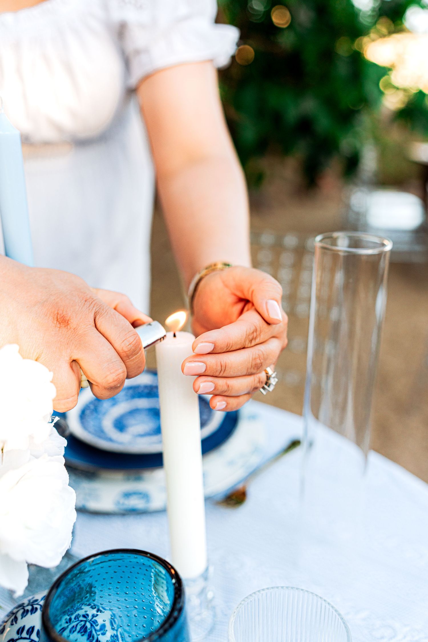 Close-up of hands lighting a white taper candle at a blue and white decorated outdoor dining table.