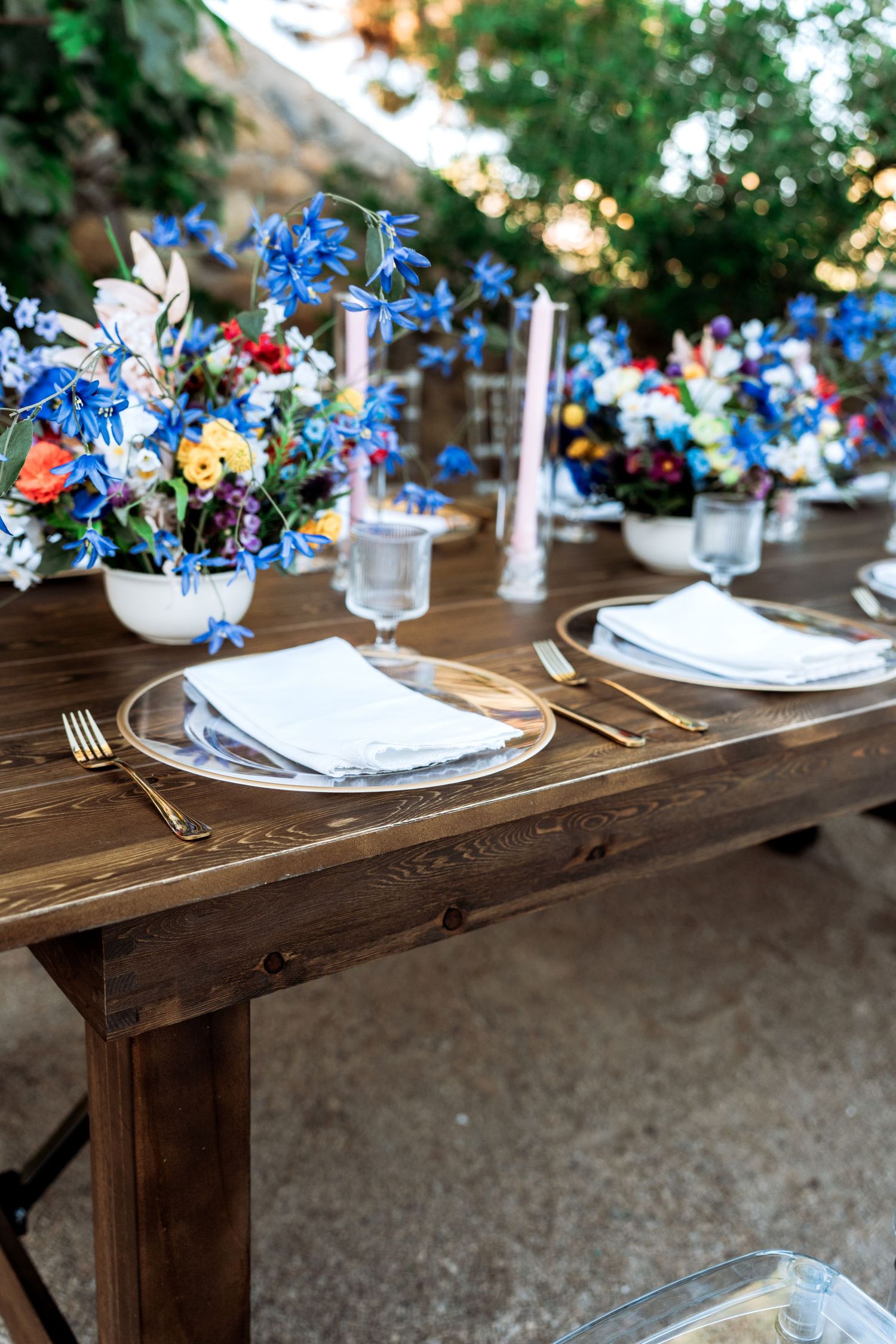 Rustic wooden reception table with gold cutlery, clear charger plates, pink candles, and blue floral arrangements.