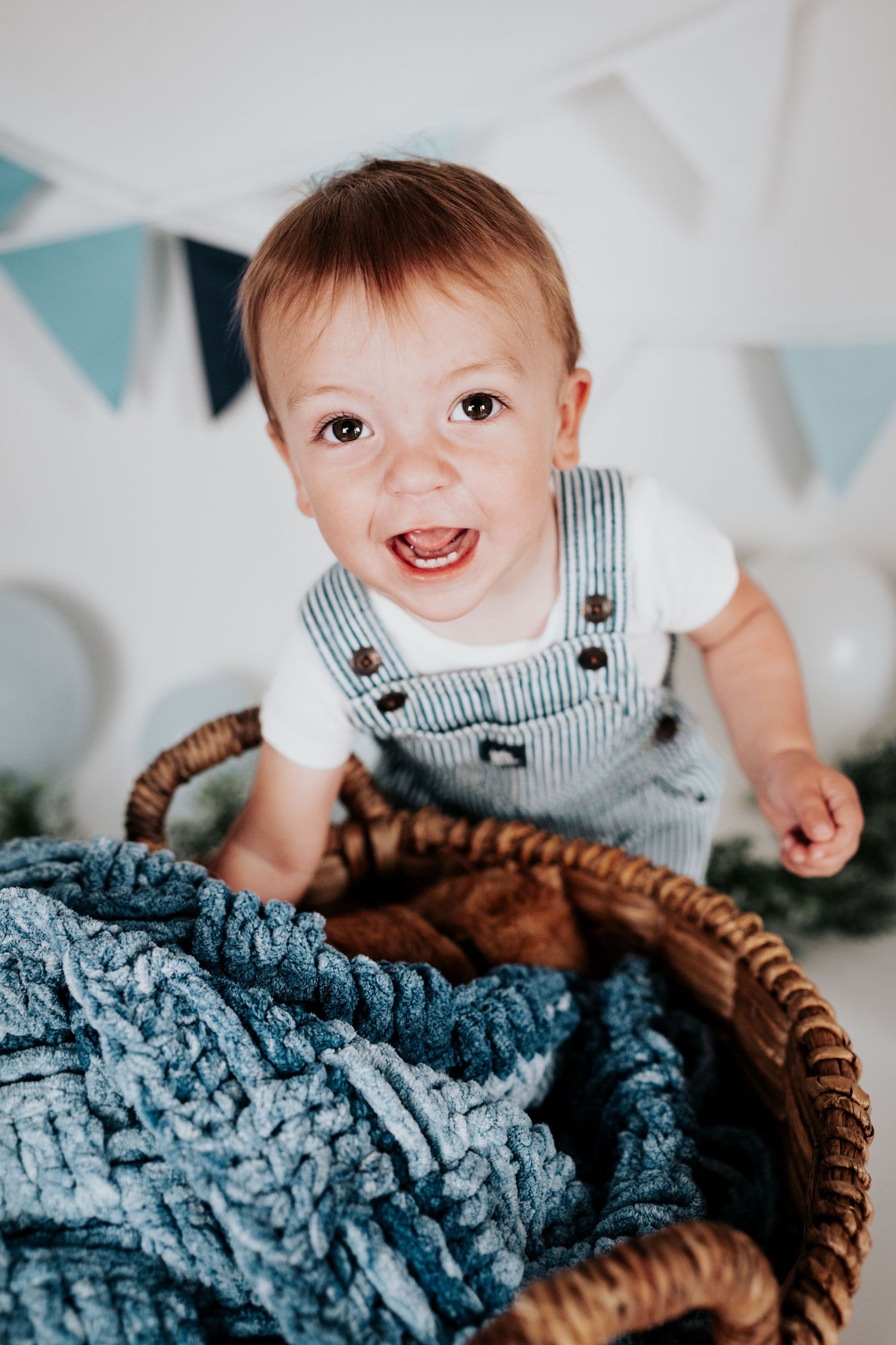 Cheerful baby boy in striped overalls leaning over a wicker basket filled with blue chunky knit blanket.