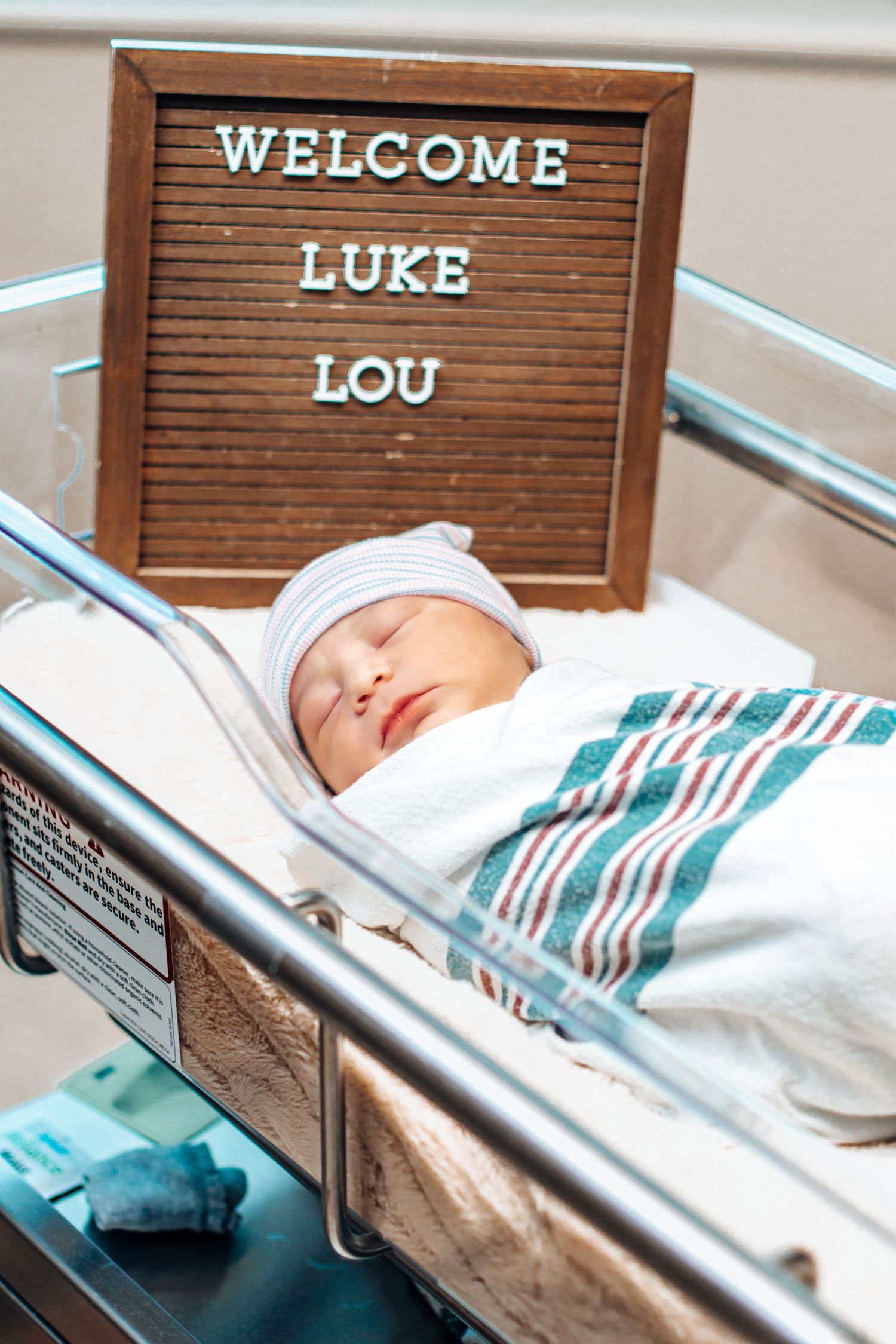 Newborn baby boy Luke Lou sleeping in hospital bassinet beside welcome letter board sign.