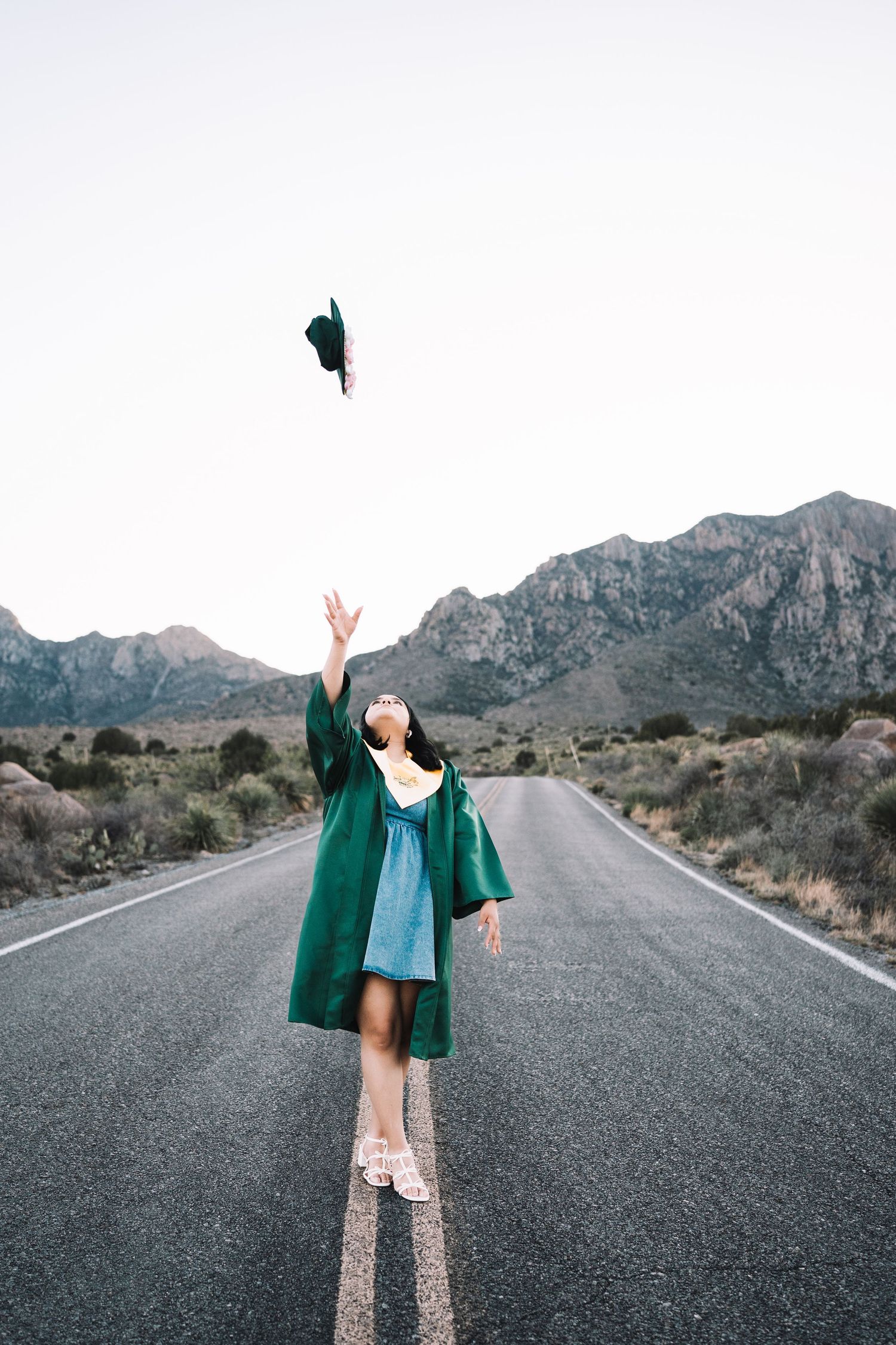 Joyful graduate in a green gown tossing her cap into the air on a scenic desert road.