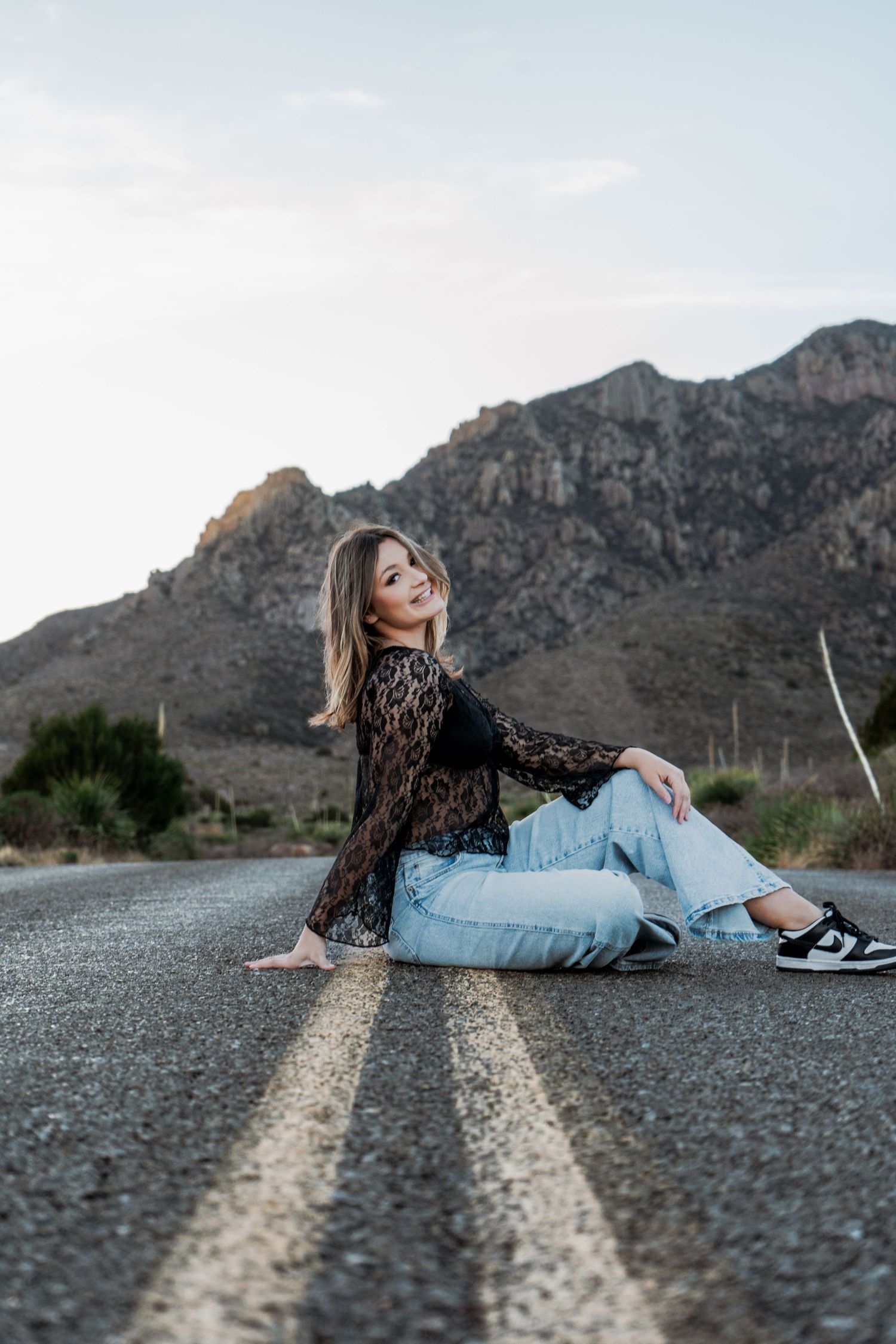 Smiling young woman in black lace top sits on mountain road centerline at golden hour.