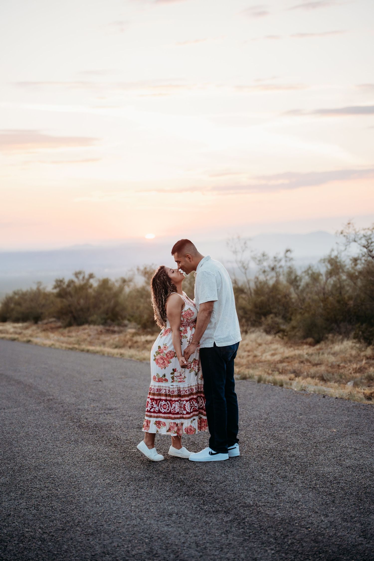 Pregnant couple sharing romantic kiss on desert road during vibrant pink and orange sunset.