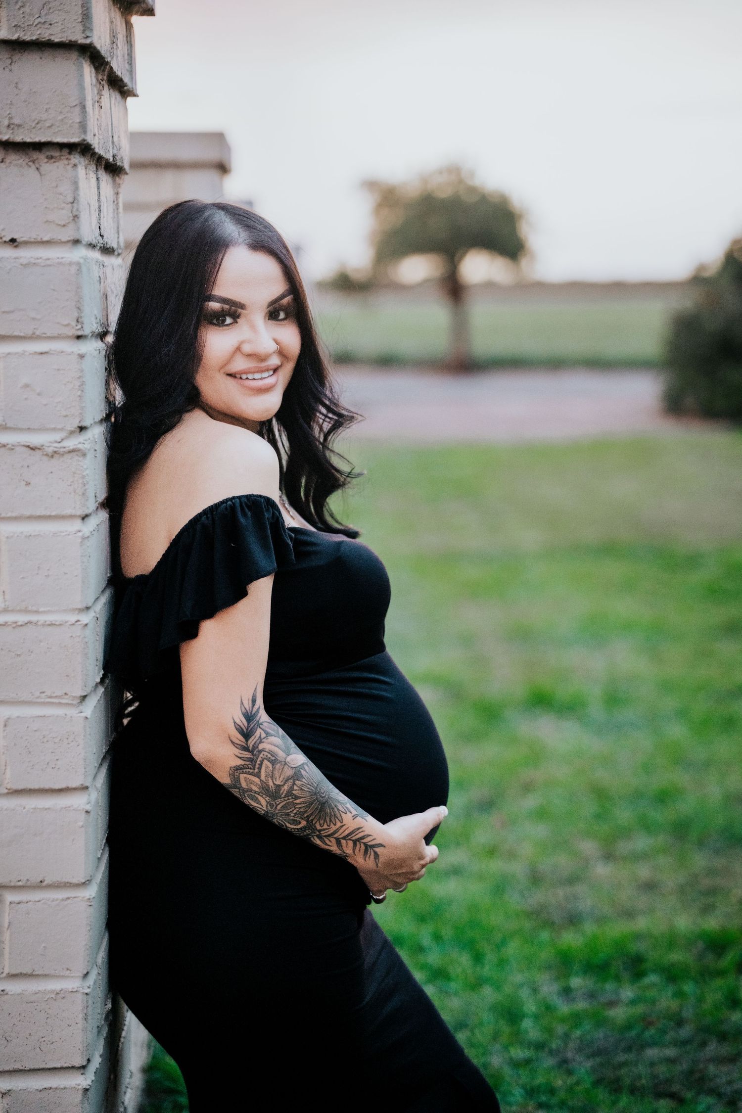 Smiling pregnant woman in black dress cradling baby bump, featuring floral arm tattoo, leaning on brick wall.