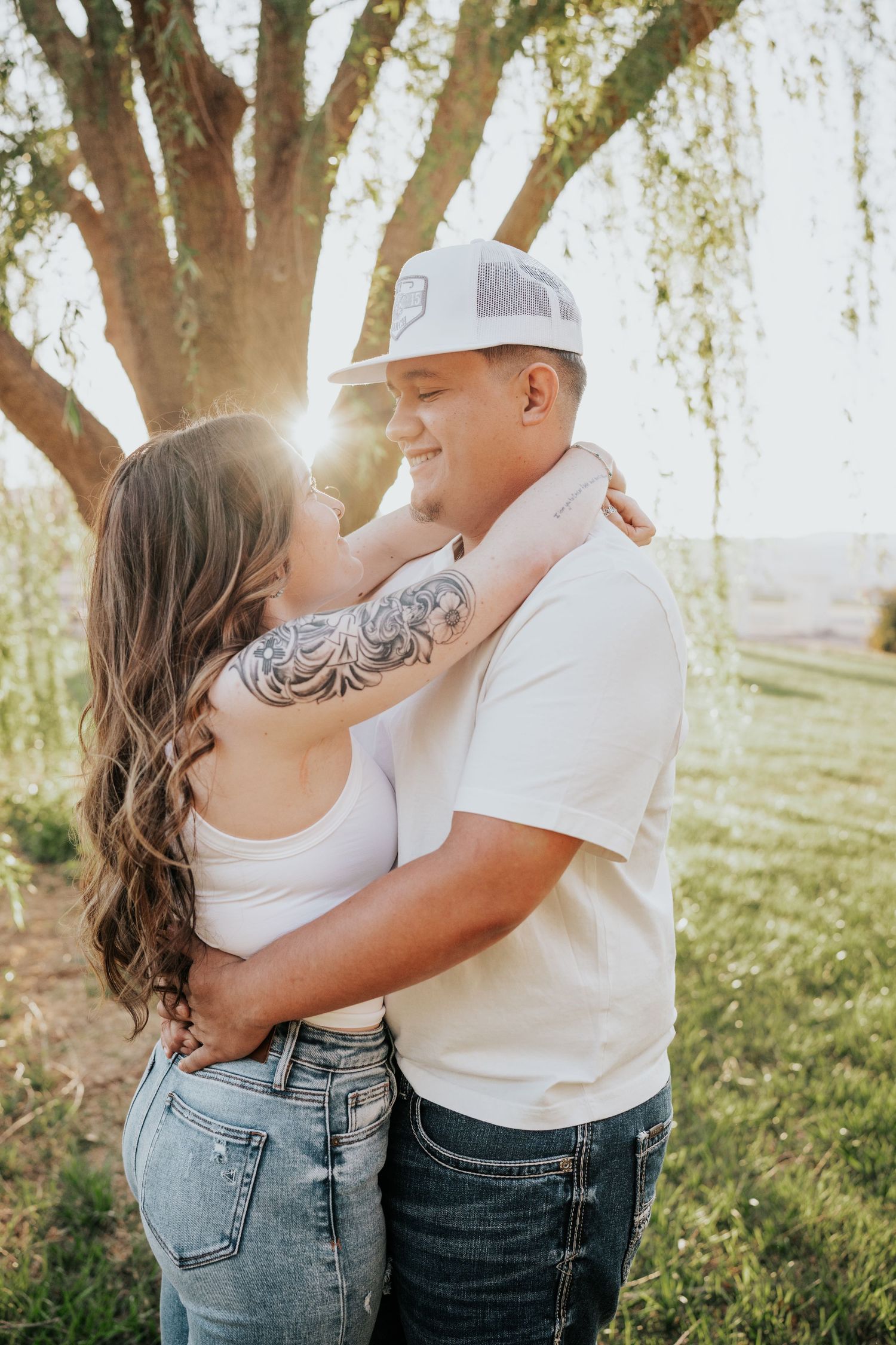 Couple embracing under tree with sun flare, woman with tattoo in white top and jeans.