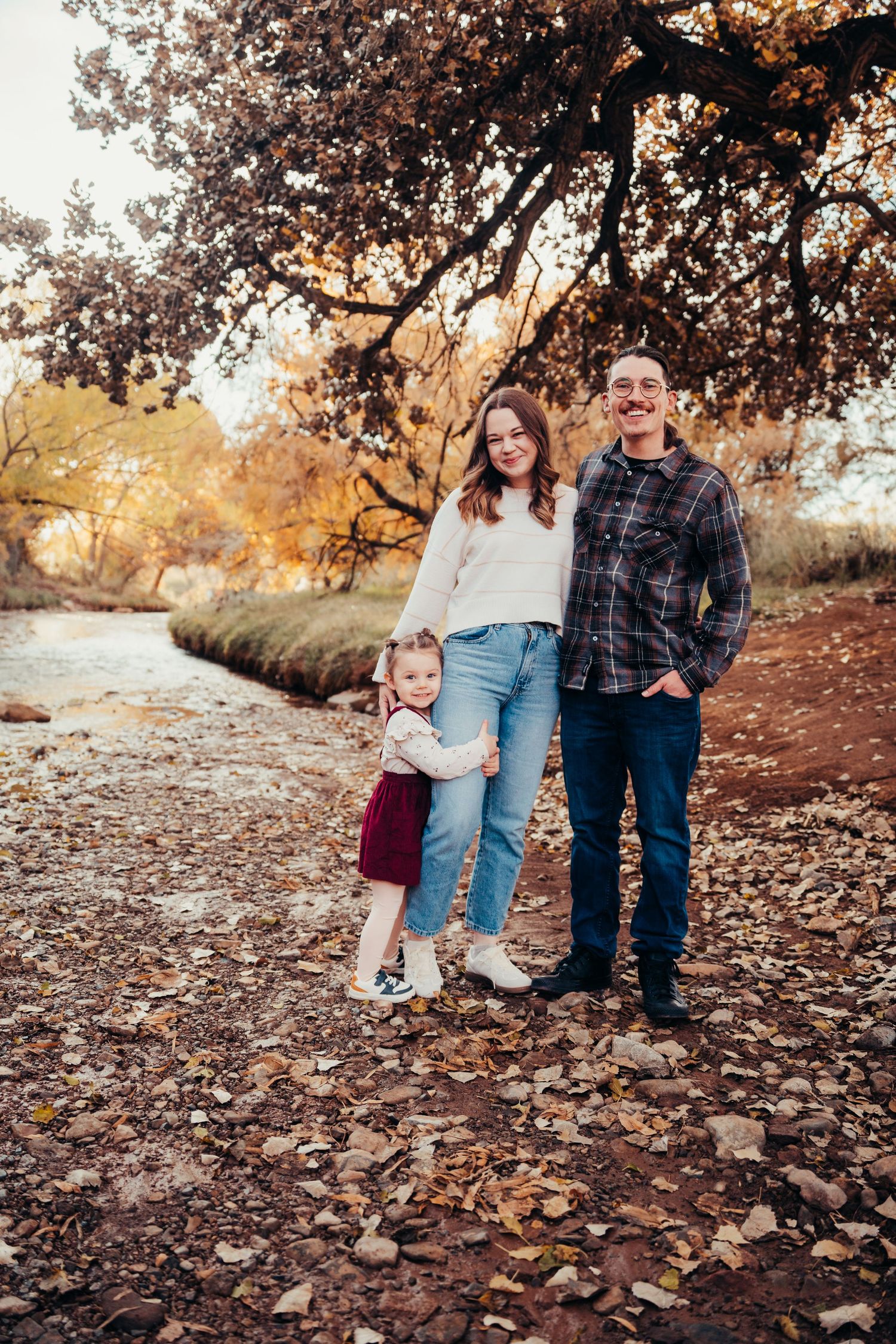 Happy family of three posing outdoors by a creek surrounded by colorful fall foliage.