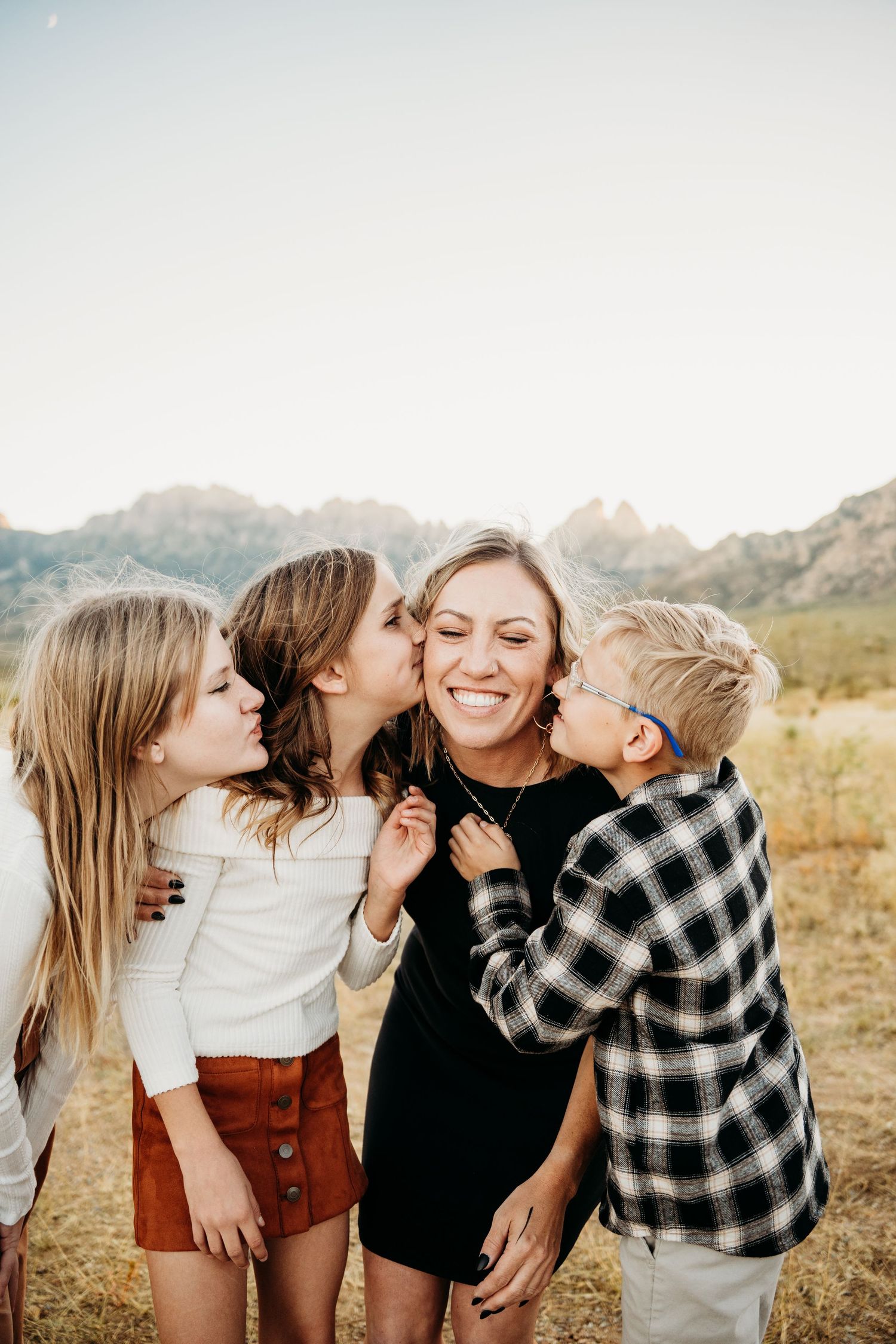 Three children lovingly kiss and hug their laughing mother outdoors in a sunny desert field.