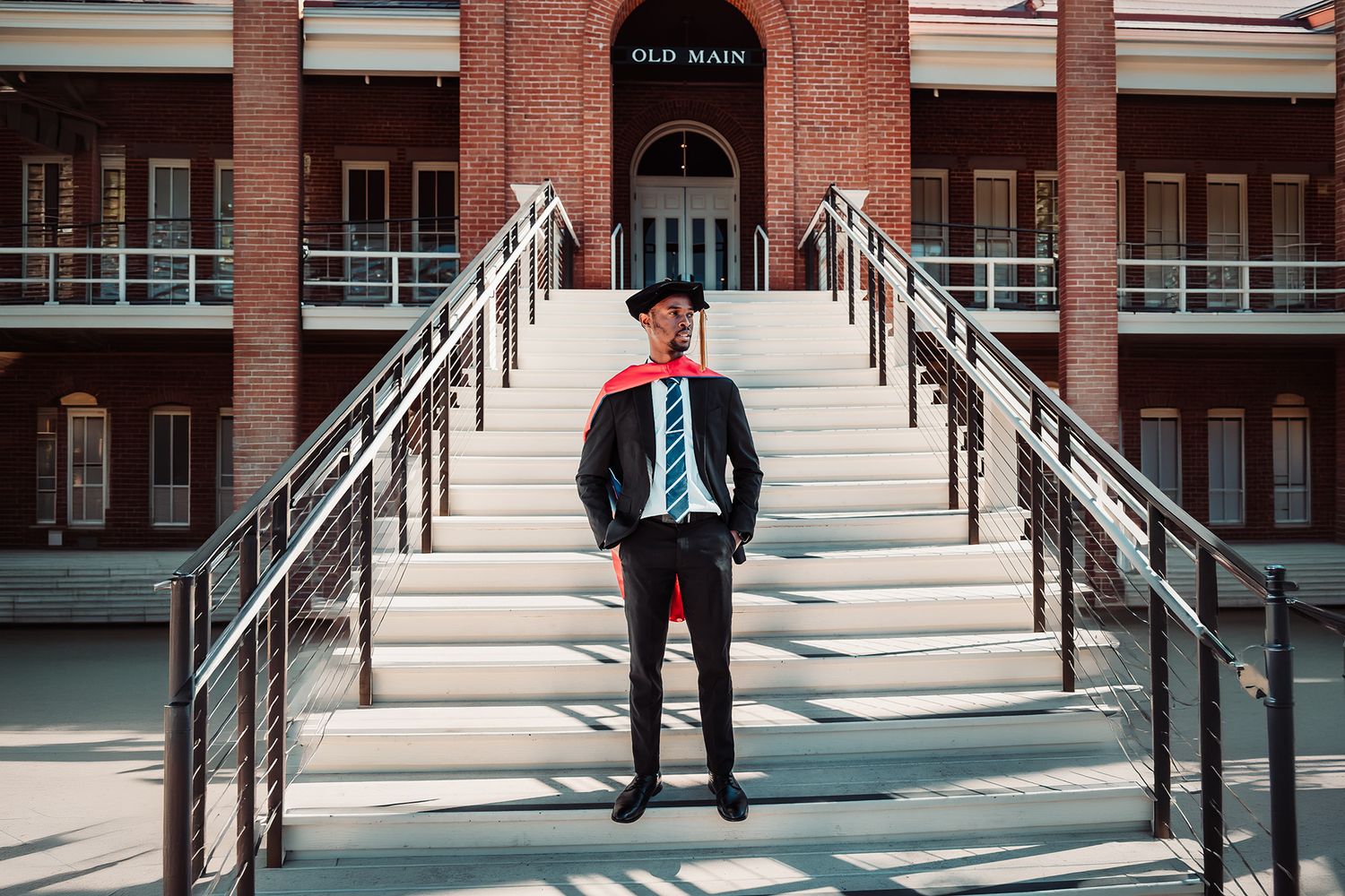 Graduate in cap and gown stands proudly on steps of Old Main building during college graduation ceremony.