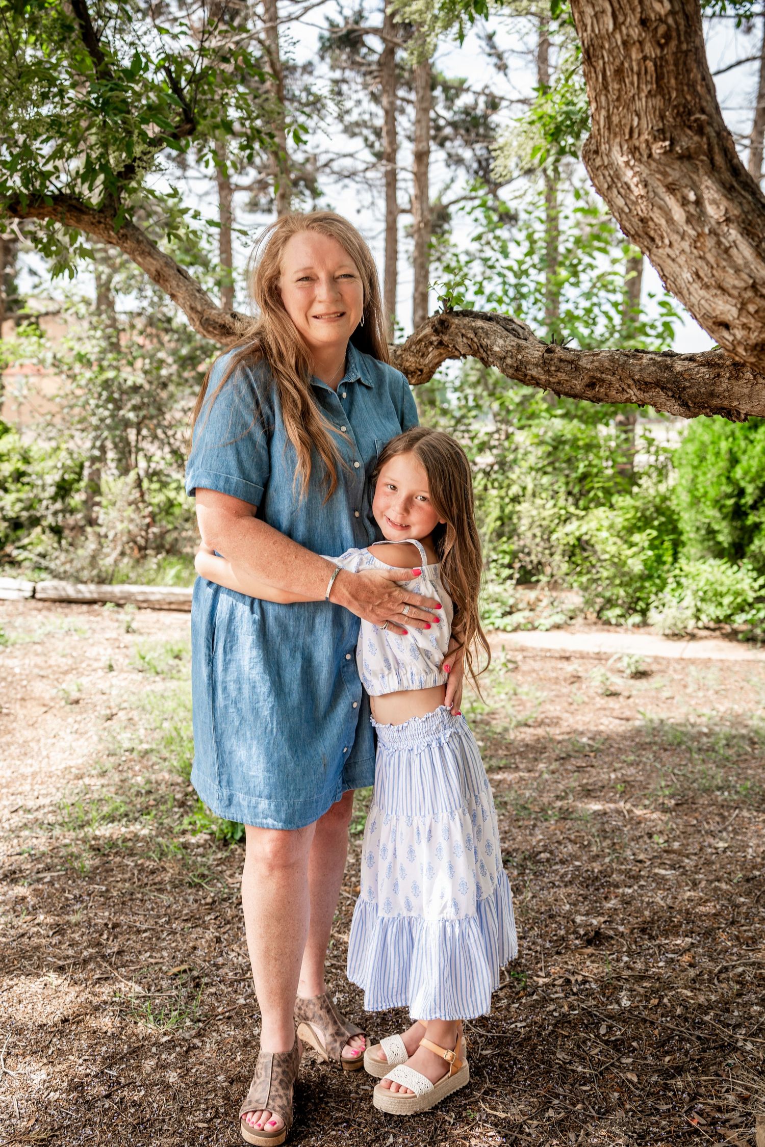 Smiling woman in blue dress posing with young girl in striped skirt under large tree.
