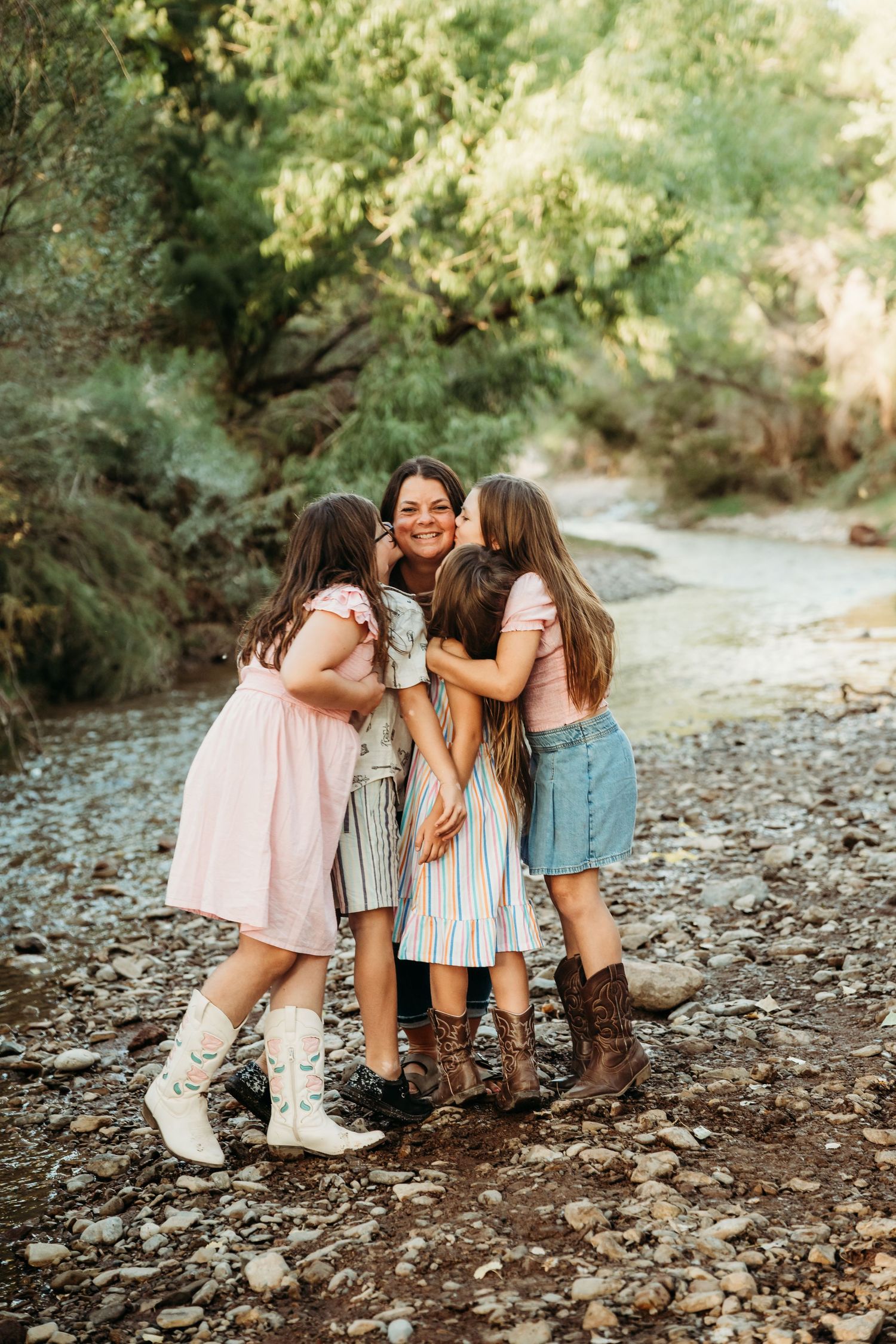 Mother being kissed on cheeks by three daughters while standing on rocky creek bed outdoors.