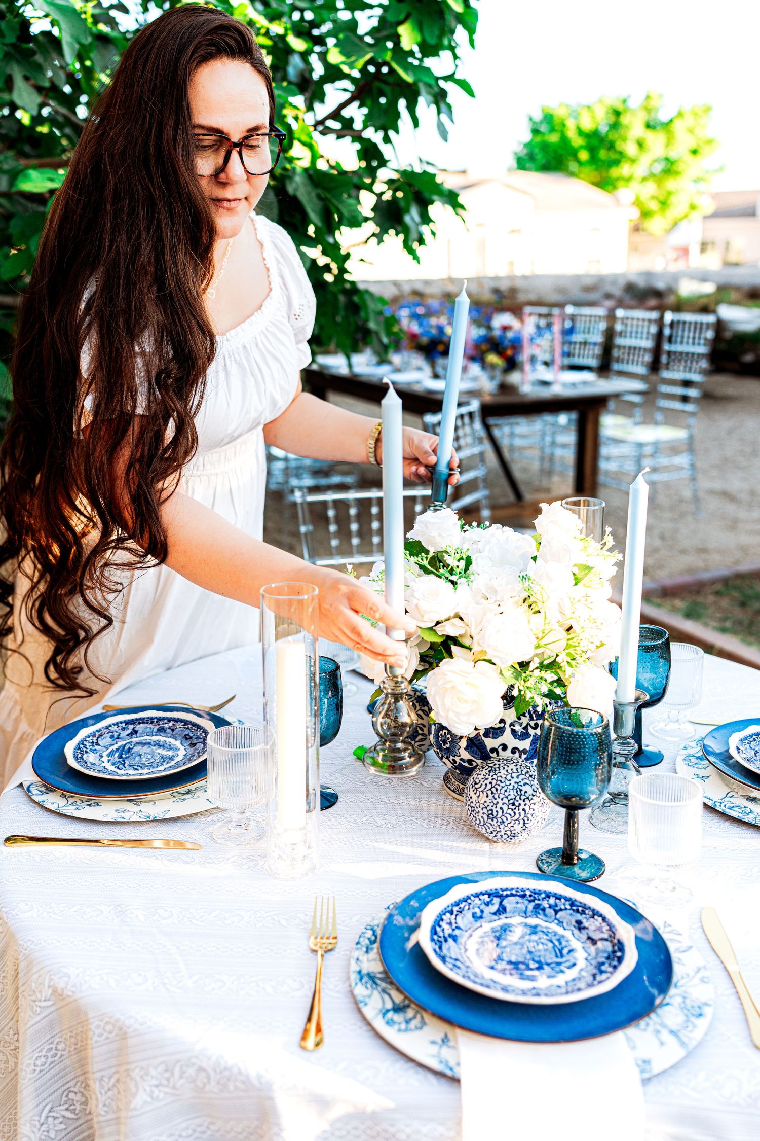 Woman in white dress arranging white rose centerpiece on blue and white china place settings at an outdoor wedding table.