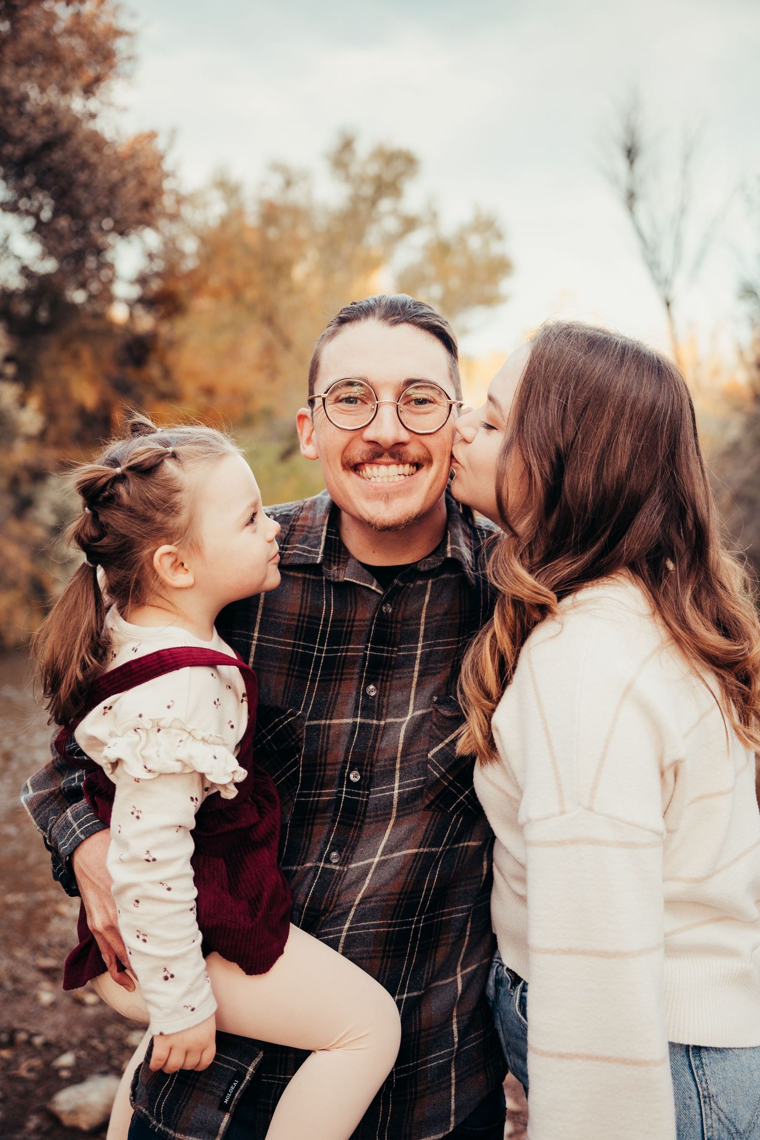 Loving family photo as mother and toddler daughter kiss father on cheeks outdoors in fall foliage setting.