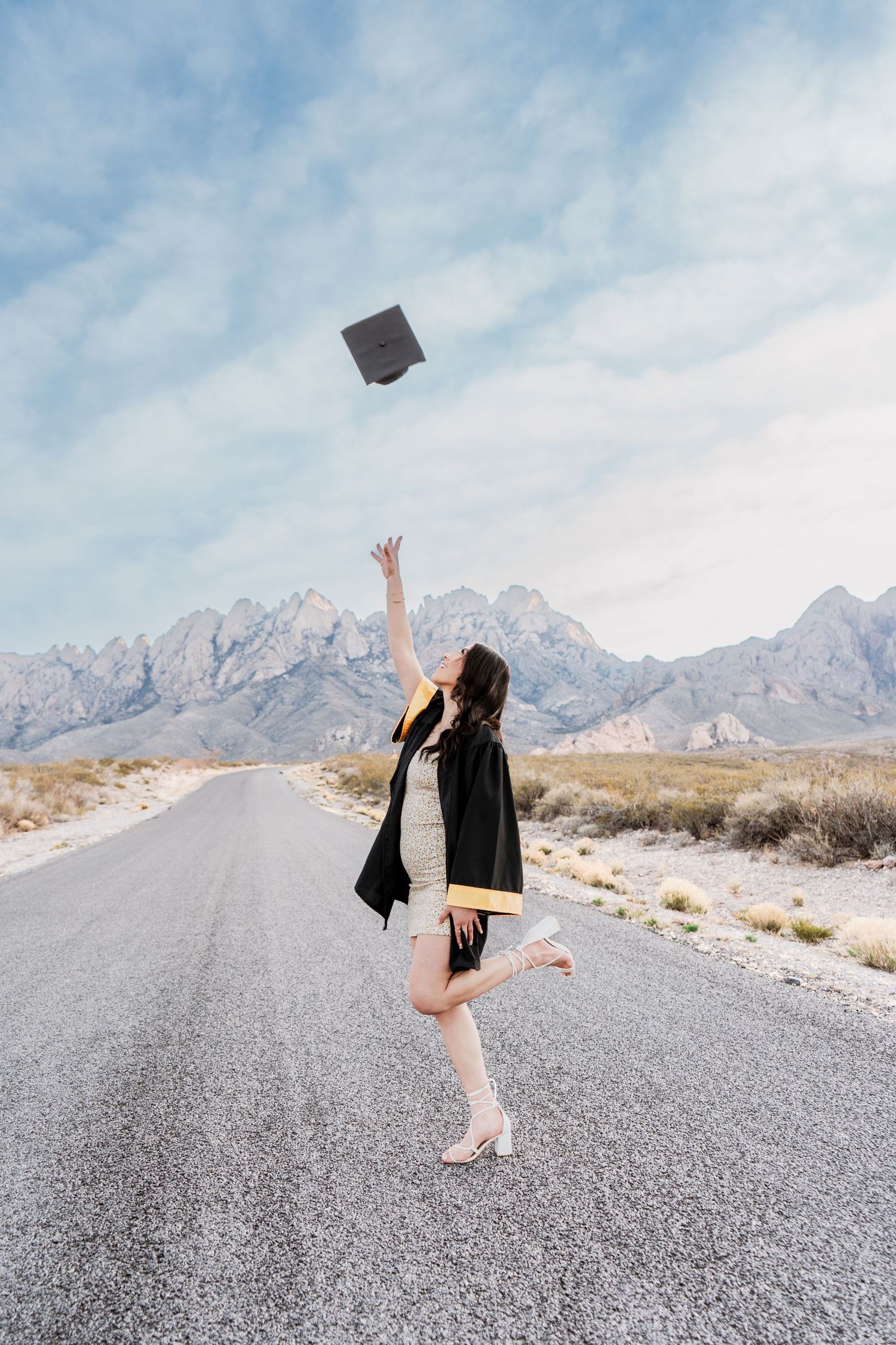 Joyful female graduate tossing cap in air on desert road with scenic mountain background.