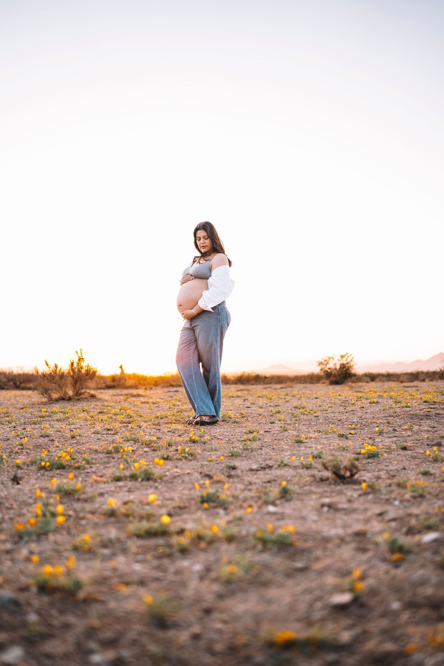 Pregnant woman cradling baby bump at golden hour sunset in desert field with wildflowers.