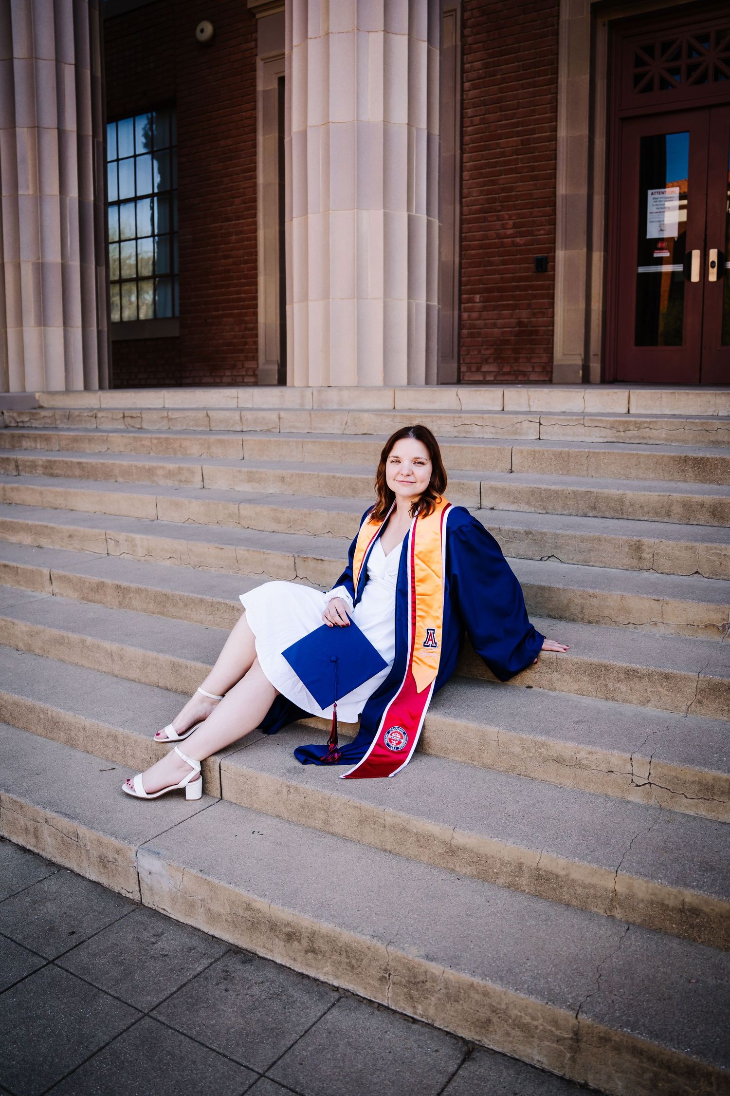 Female graduate sits on stone steps in cap and gown with diploma at university building.