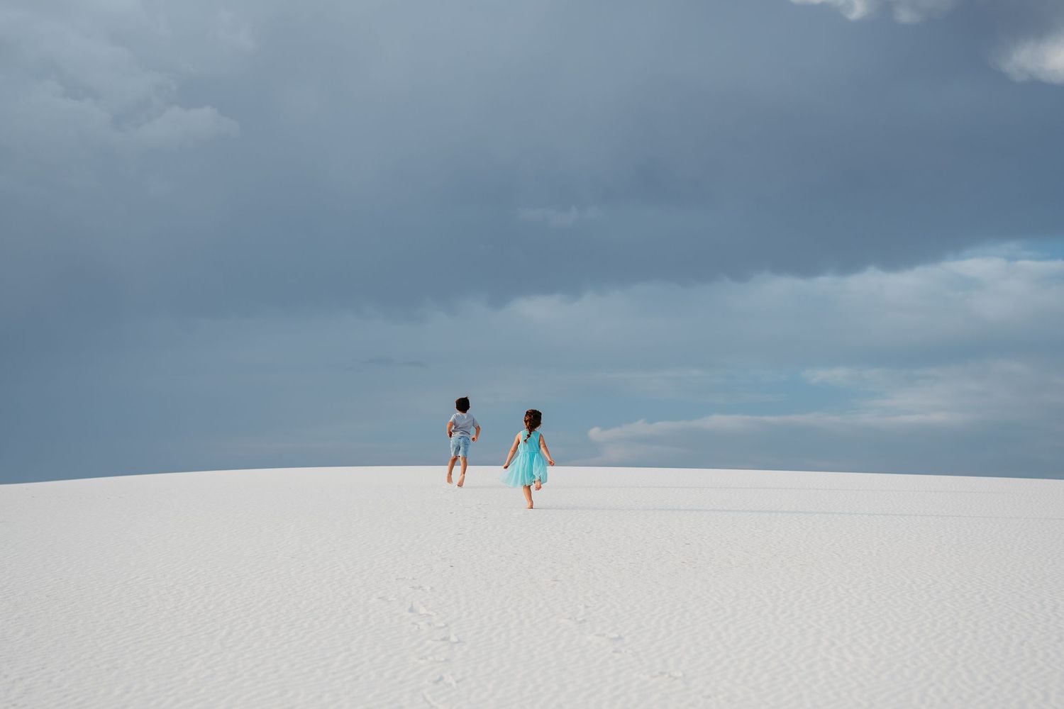 Two children in blue outfits walk across vast white sand dunes under a dramatic cloudy sky.