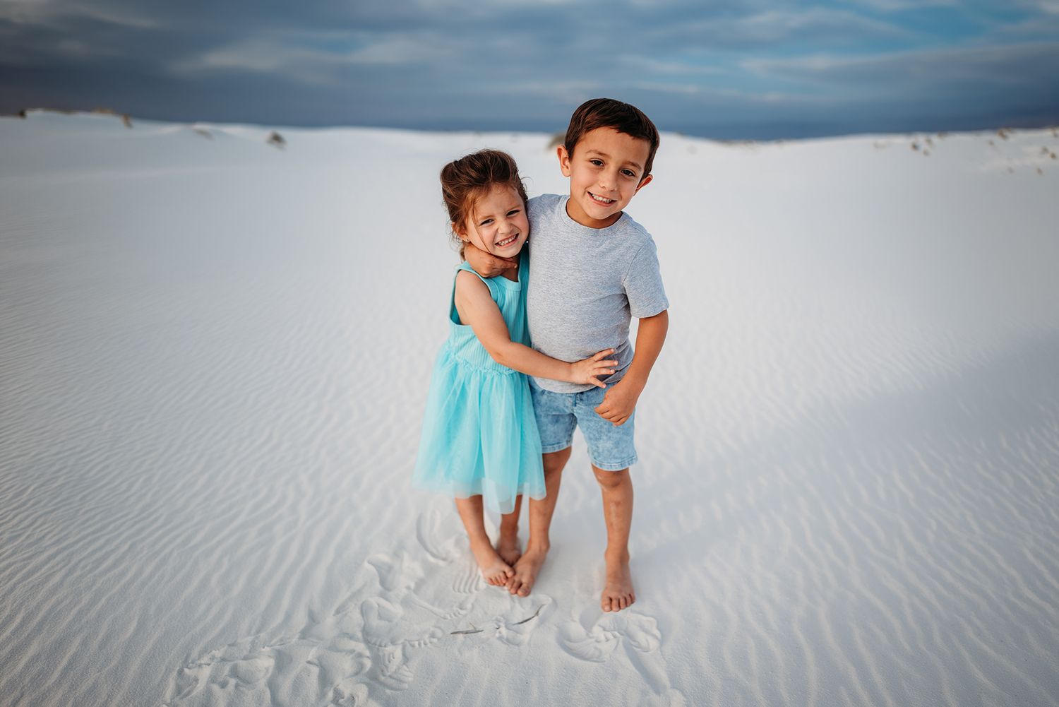 Two young siblings hugging and smiling together on white sand dunes under a dramatic cloudy sky.
