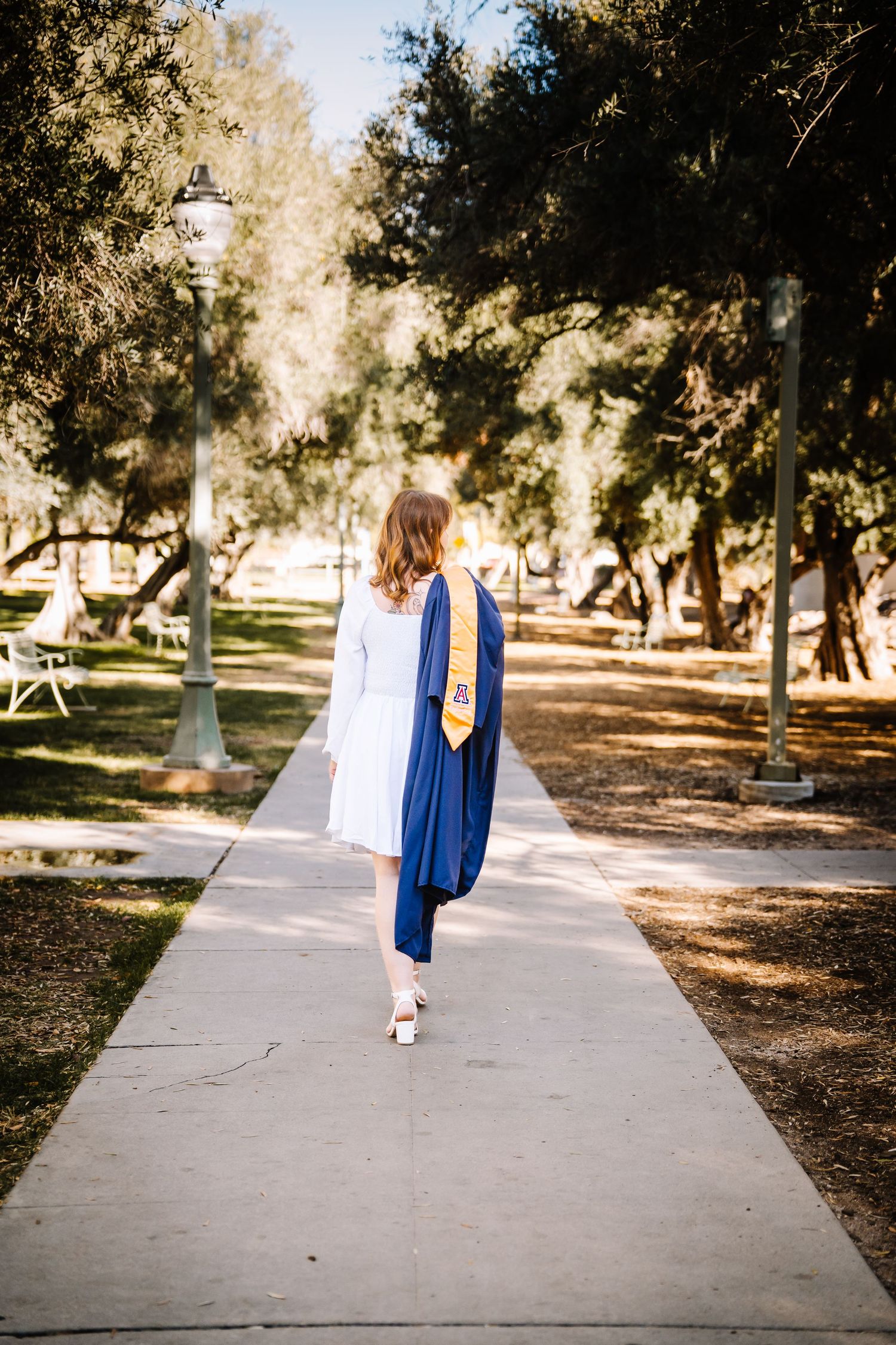 A female graduate in navy blue gown walks away down a tree-lined campus pathway wearing white heels.