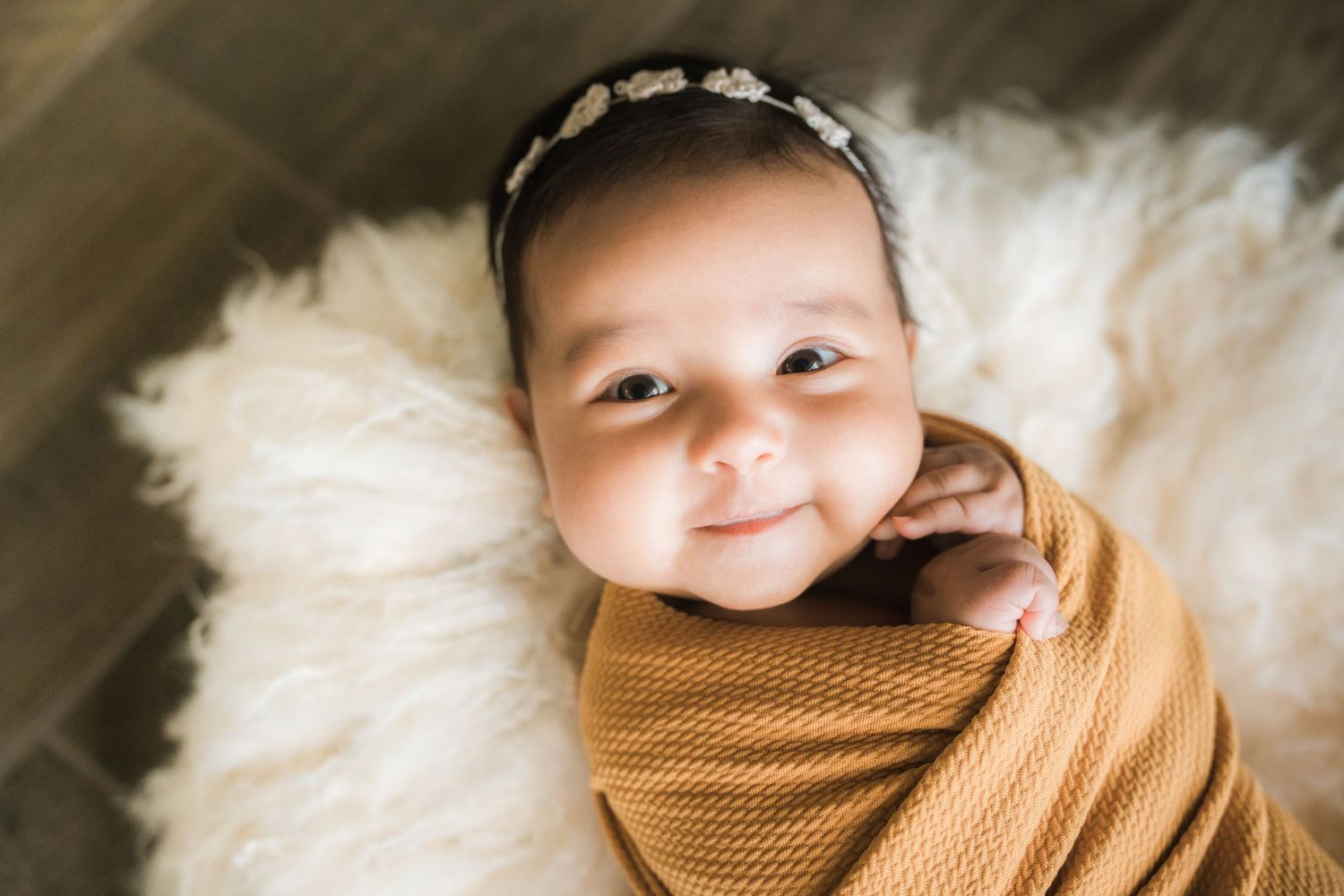 Smiling awake newborn baby wrapped in mustard yellow swaddle with daisy headband on white fluffy blanket.