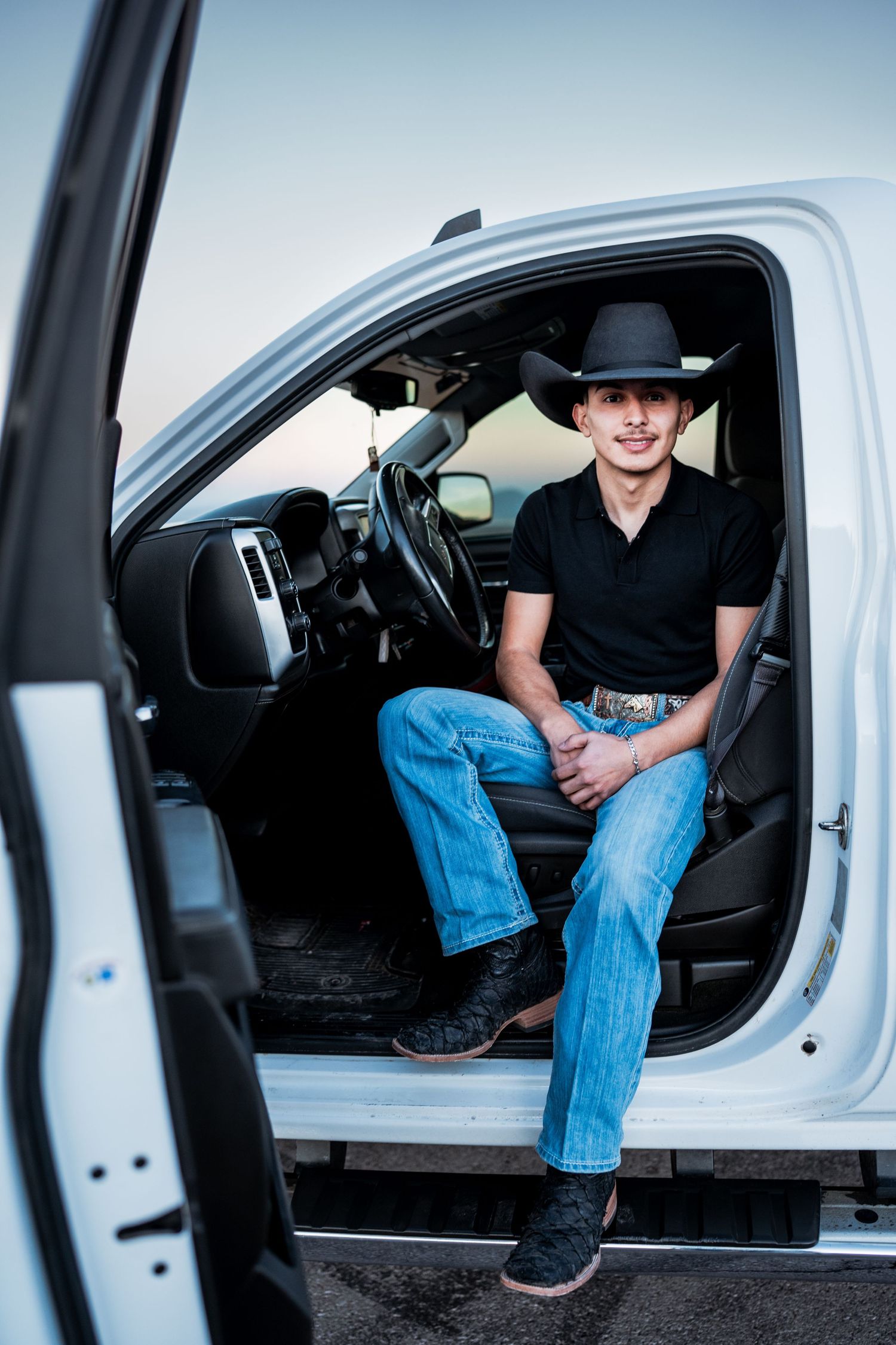 Smiling young cowboy in black hat sits in driver seat of white pickup truck with door open.