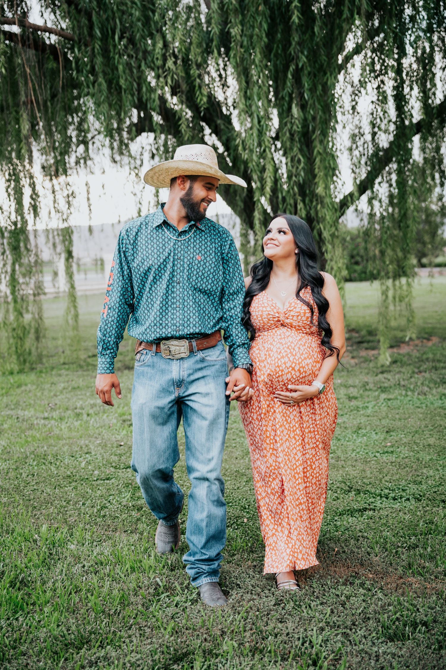 Pregnant couple walking hand in hand on green grass under weeping willow, man in cowboy hat and teal shirt.