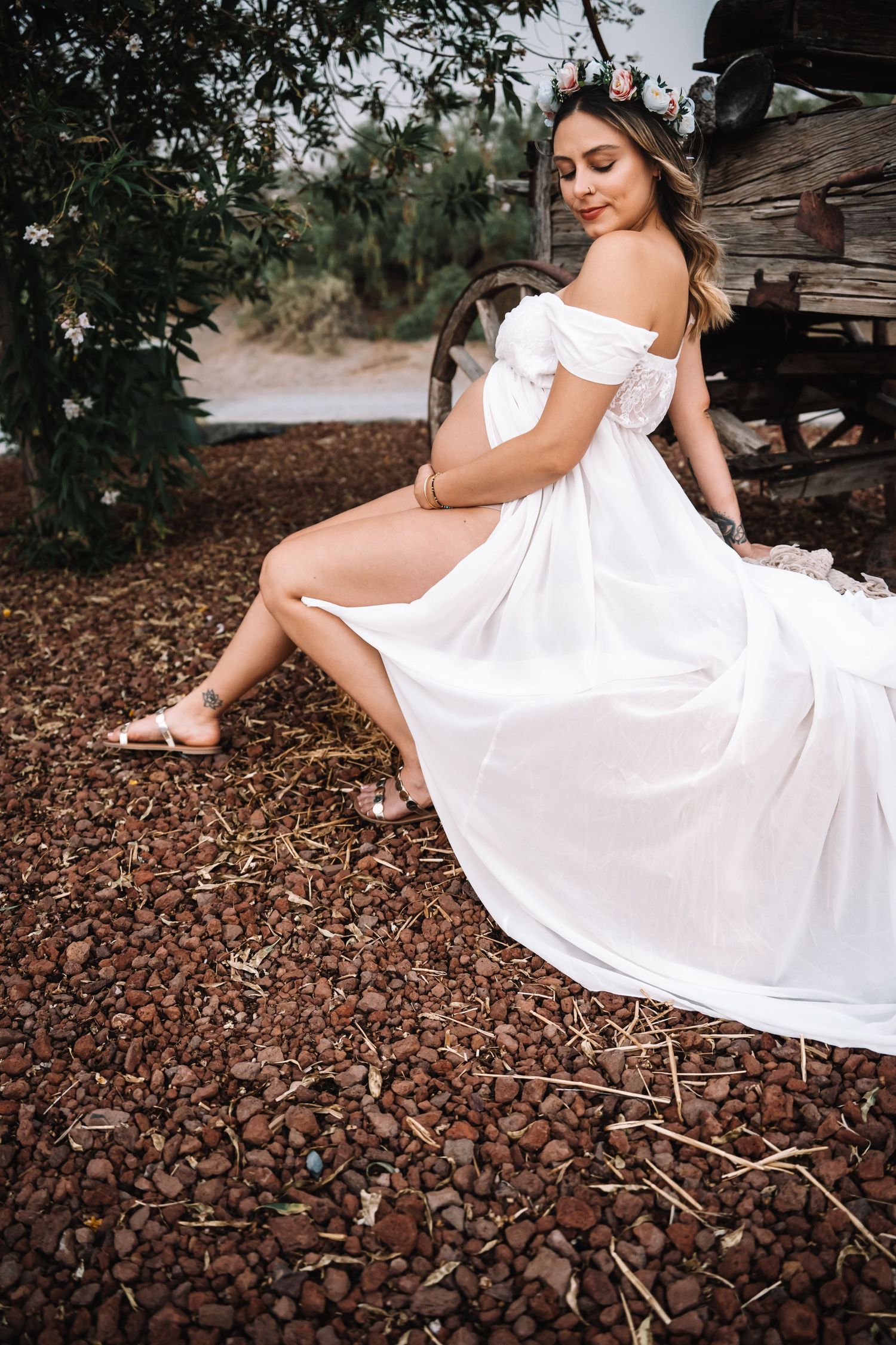 Pregnant woman in elegant white off-shoulder dress with floral crown sits on rustic ground outdoors.