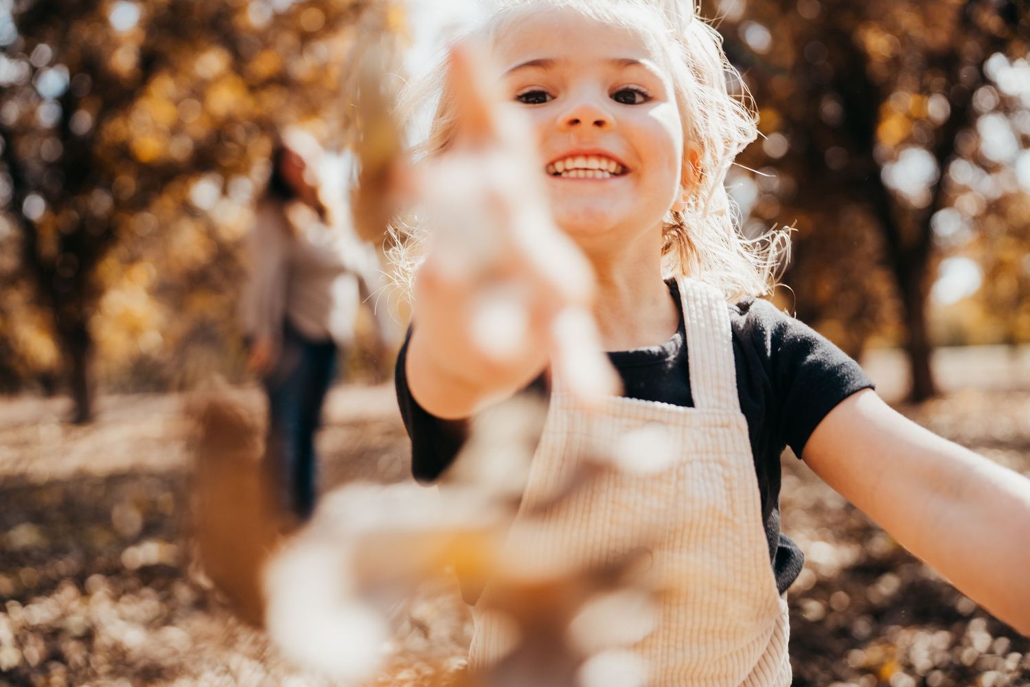 Smiling blonde girl in overalls reaching toward camera with autumn leaves in background.